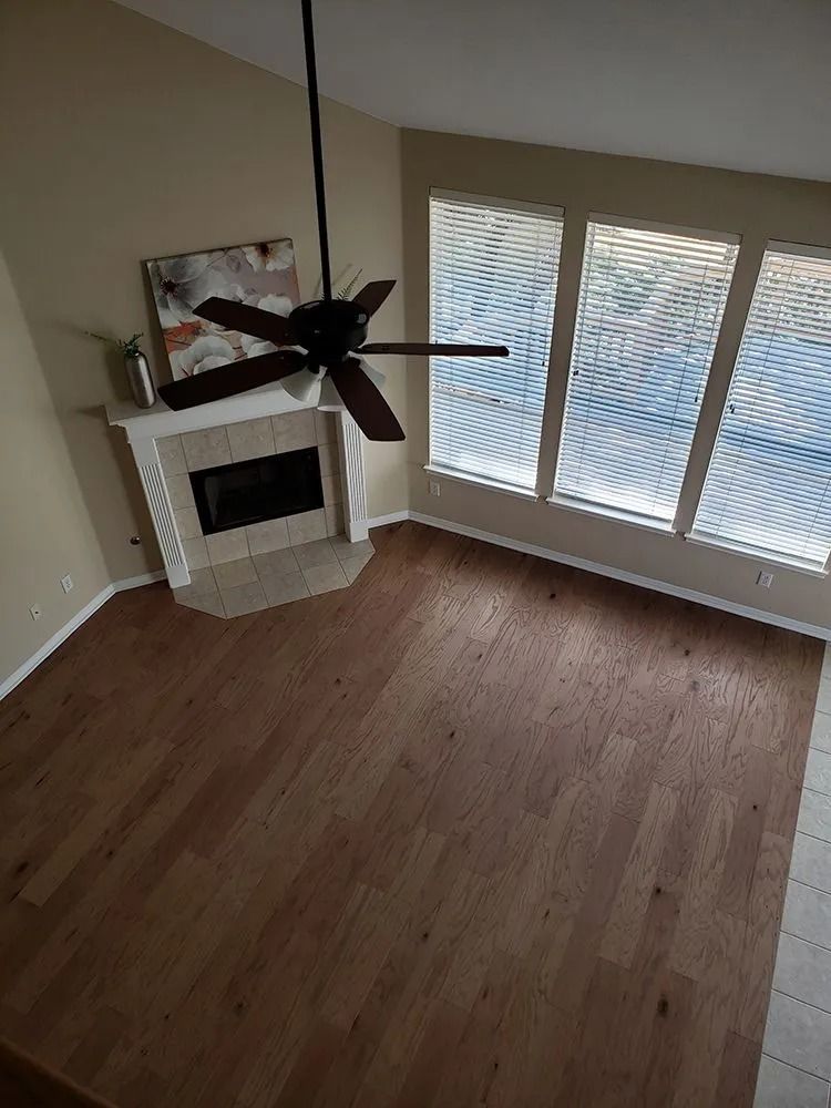 Empty living room with hardwood floors, fireplace, ceiling fan, and large windows with blinds