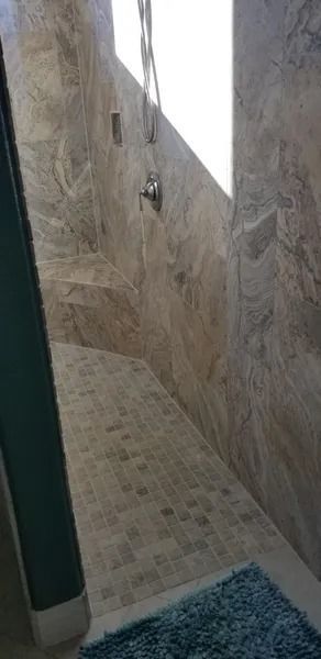 Narrow tiled shower stall with beige stone walls and blue bath mat at the entrance