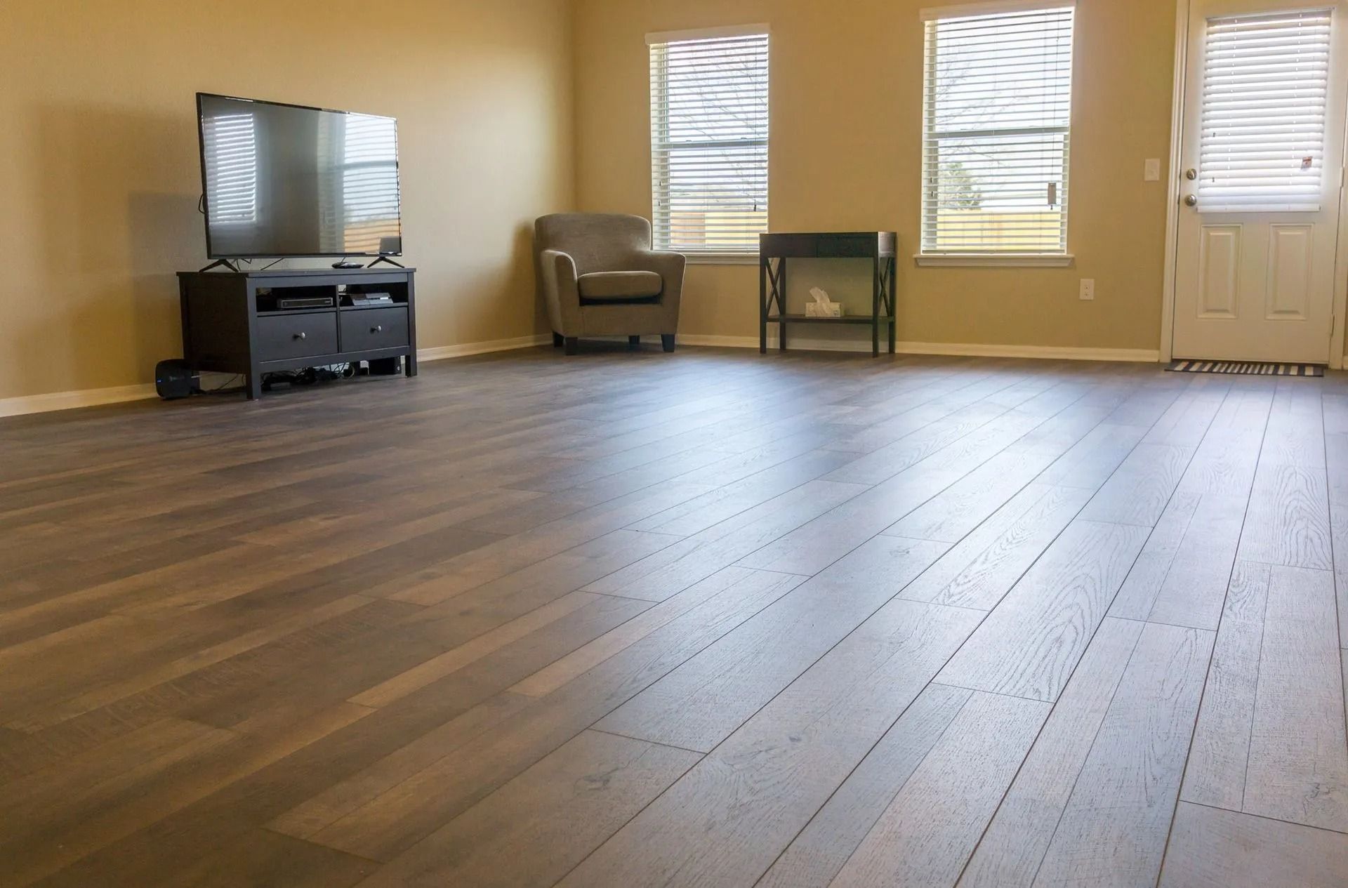 Empty living room with hardwood floors, TV stand, armchair, table, windows, and a door with blinds