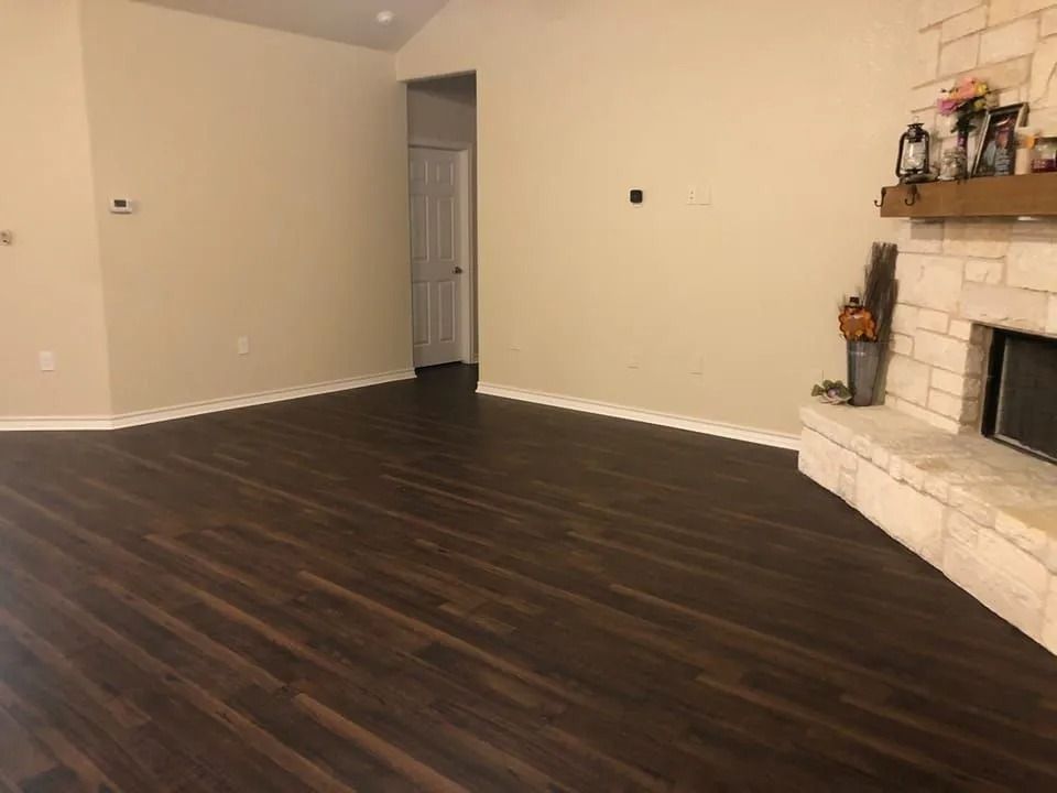 Empty living room with dark wood floors, beige walls, and a stone fireplace on the right