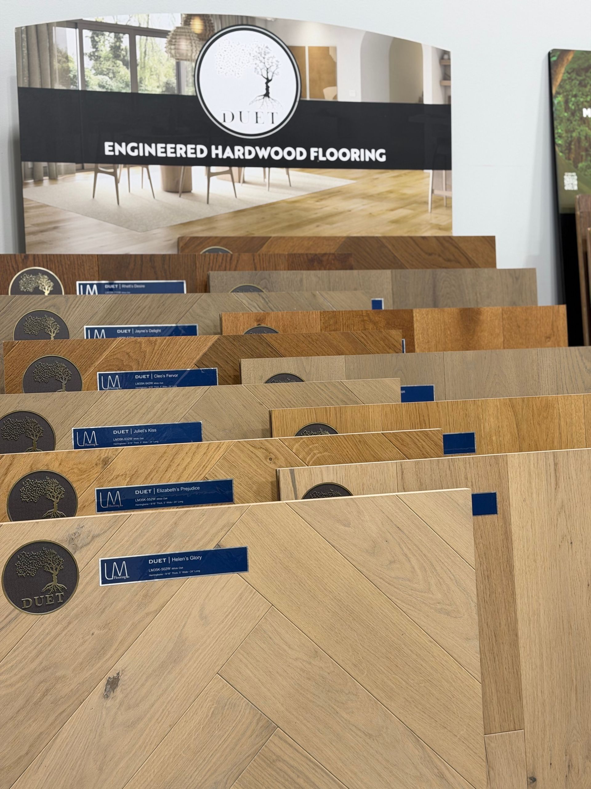Display of engineered hardwood flooring samples in tan, brown, and gray tones with a product brochure above