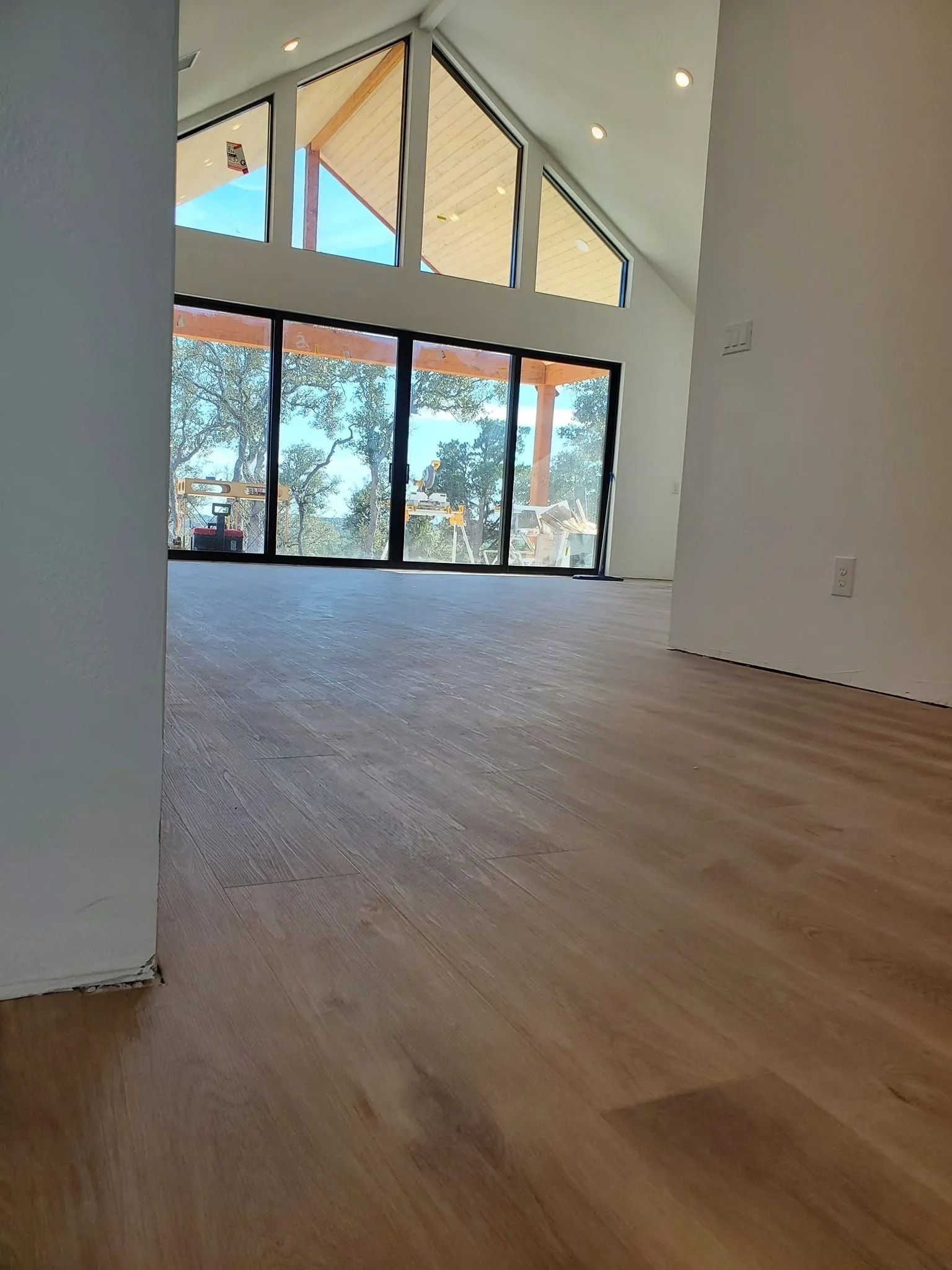 Empty modern room with light wood floors, high slanted ceiling, and large windows overlooking trees