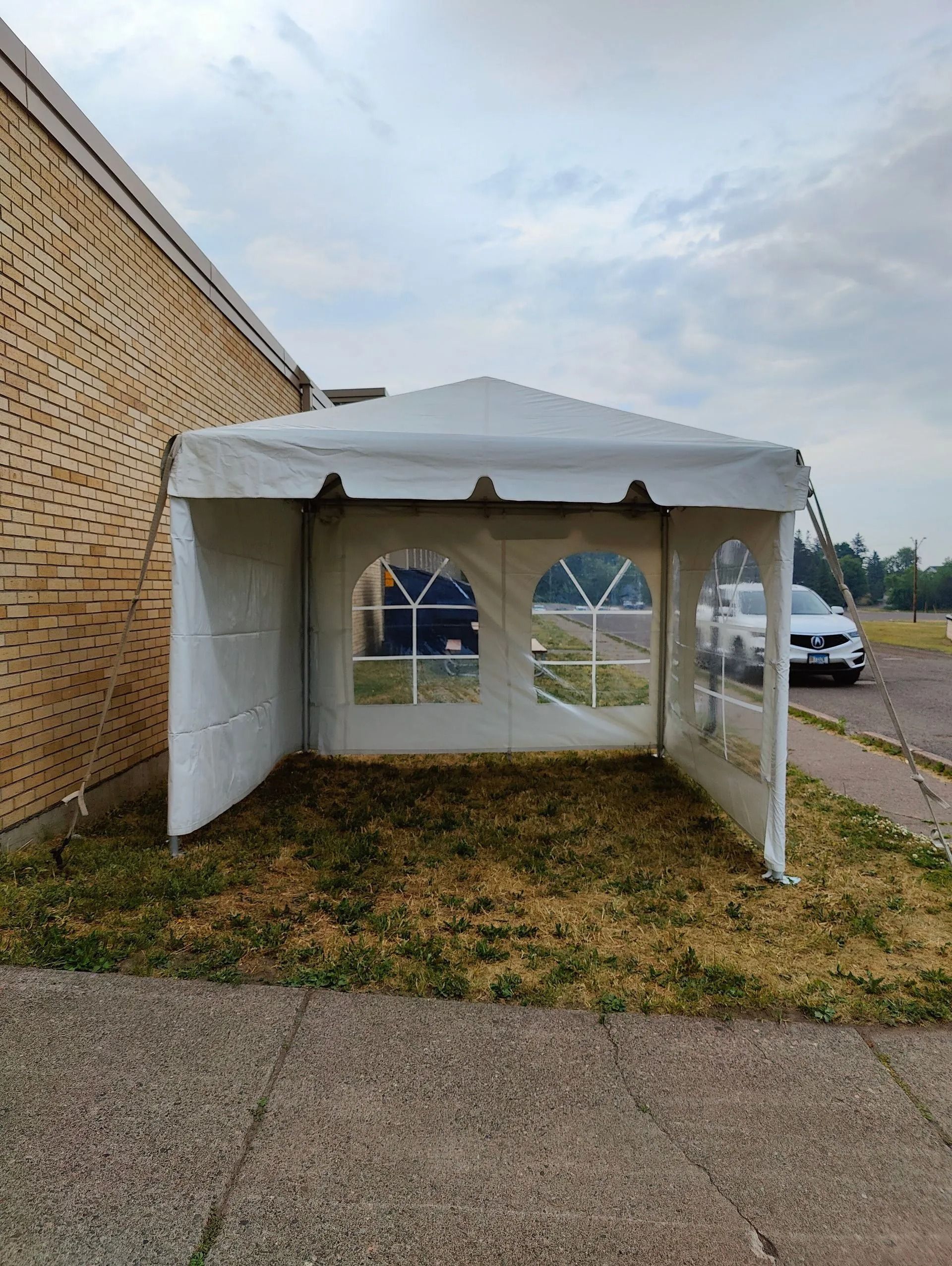 White tent with clear window panels sits on dry grass next to a brick building.