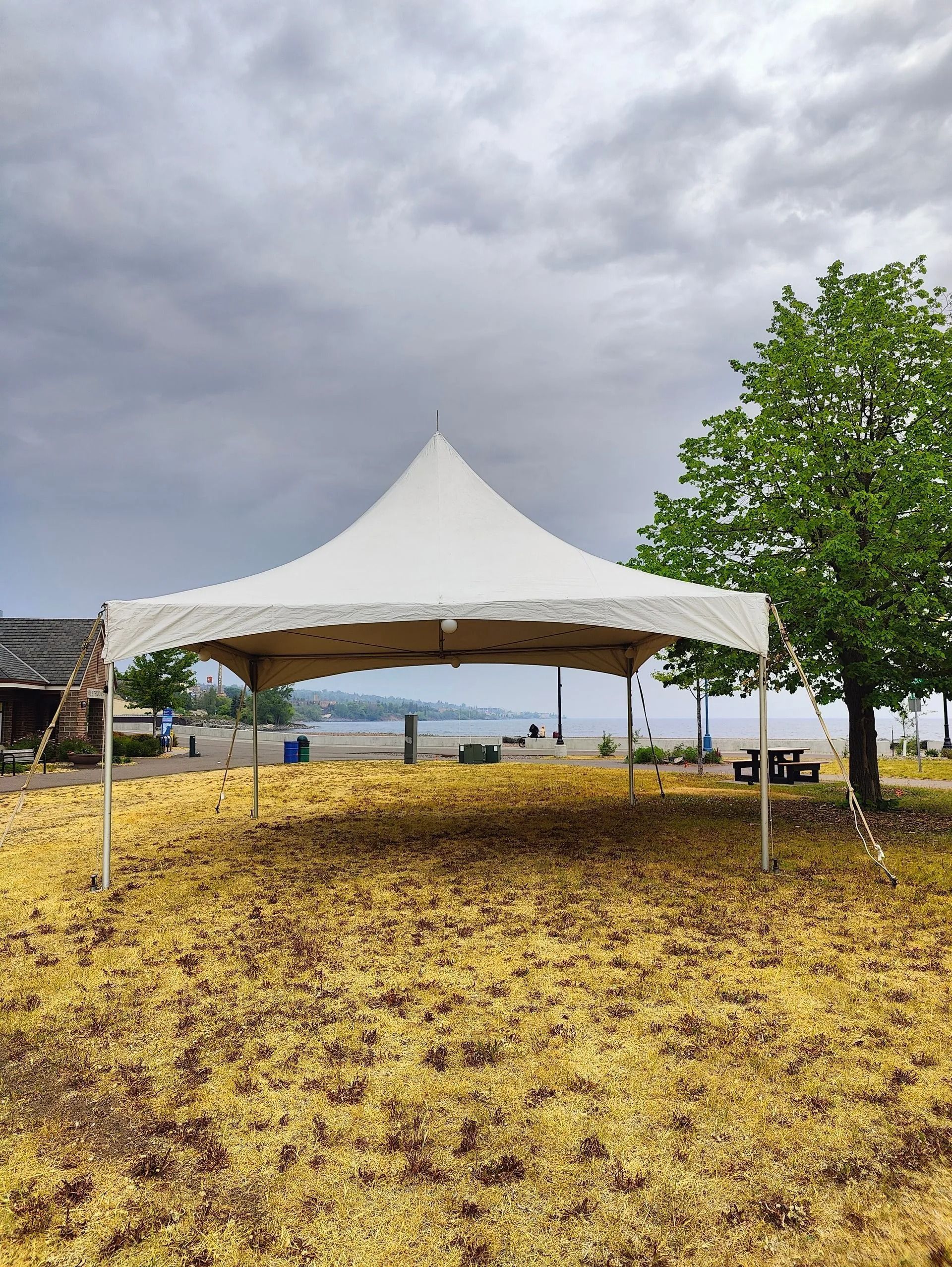 White tent on a lawn by a beach under cloudy skies, with a tree in the background.