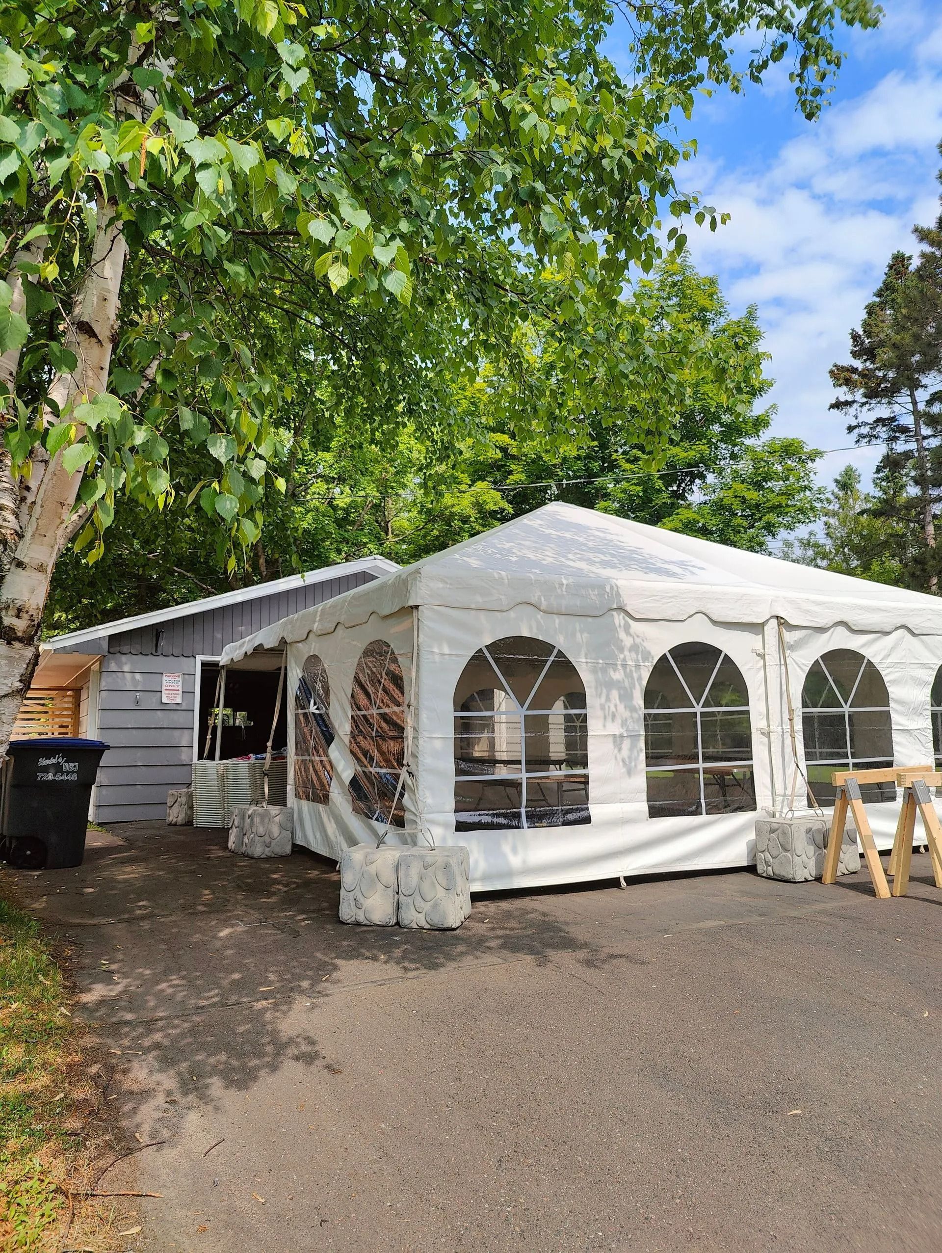 White tent beside a small gray building, set on a gravel driveway, surrounded by trees.