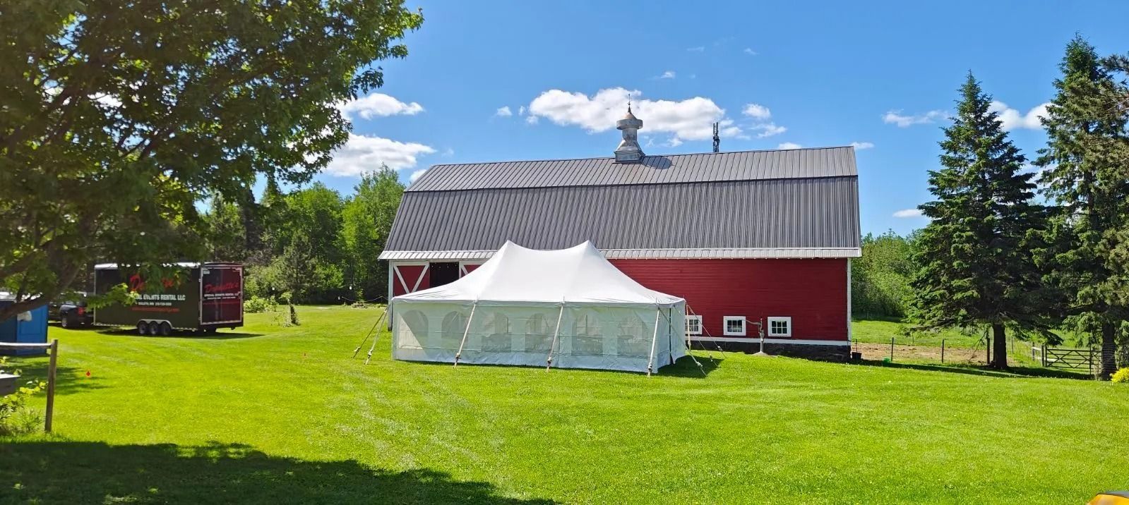 Red barn and white tent on a green lawn under a blue sky with trees.