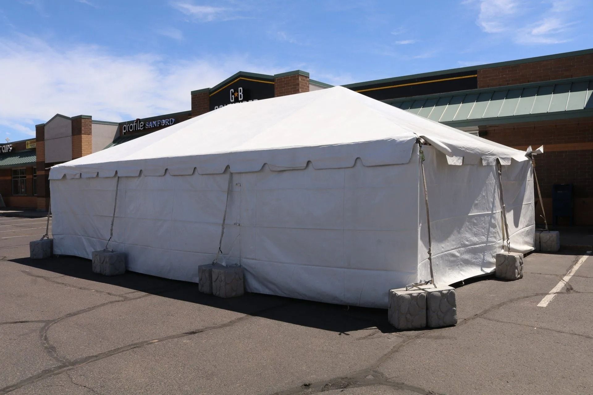 White tent set up in a parking lot, concrete blocks at its base. A building is in the background.