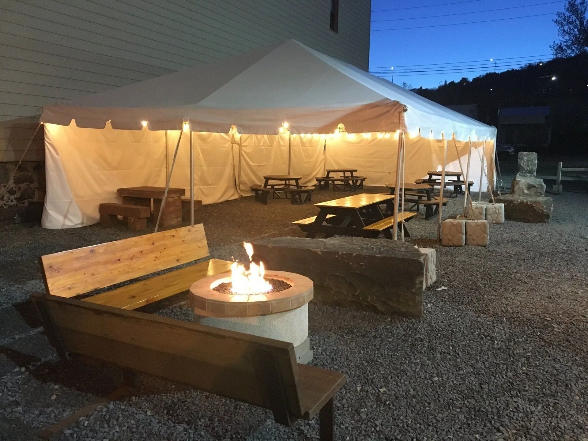 An outdoor area with a tent, picnic tables, benches, a fire pit, and string lights. Nighttime setting.