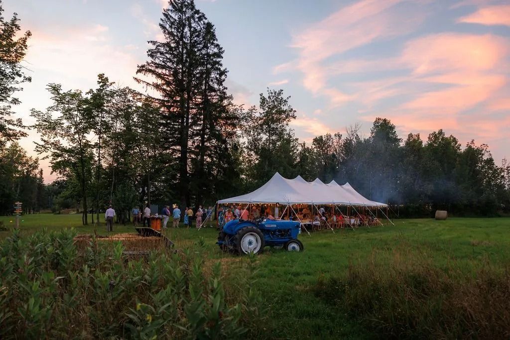 Blue tractor in field near white tent with people gathered; sunset sky.