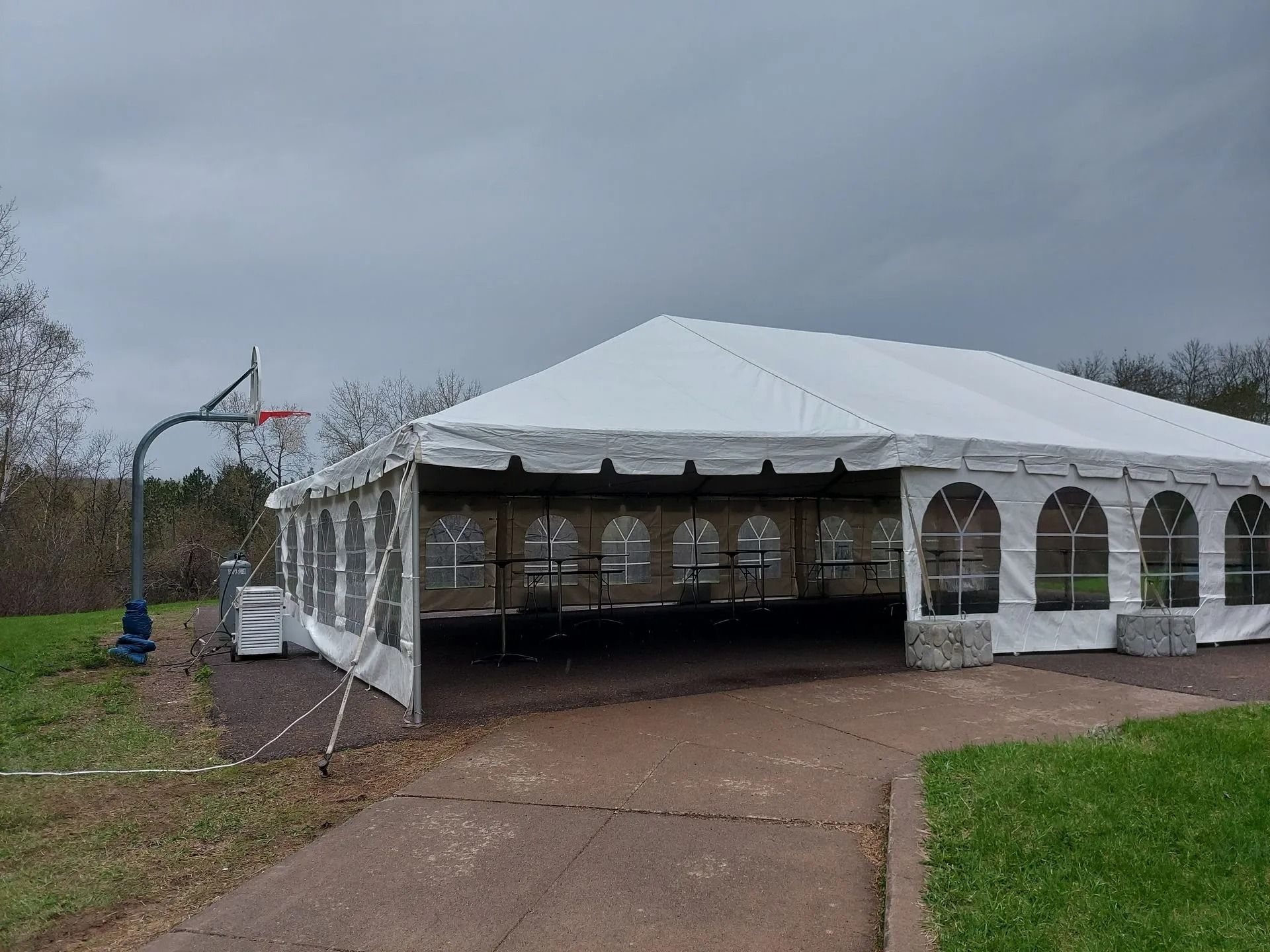 White tent with clear side panels, on a paved area, basketball hoop to the left, overcast sky.