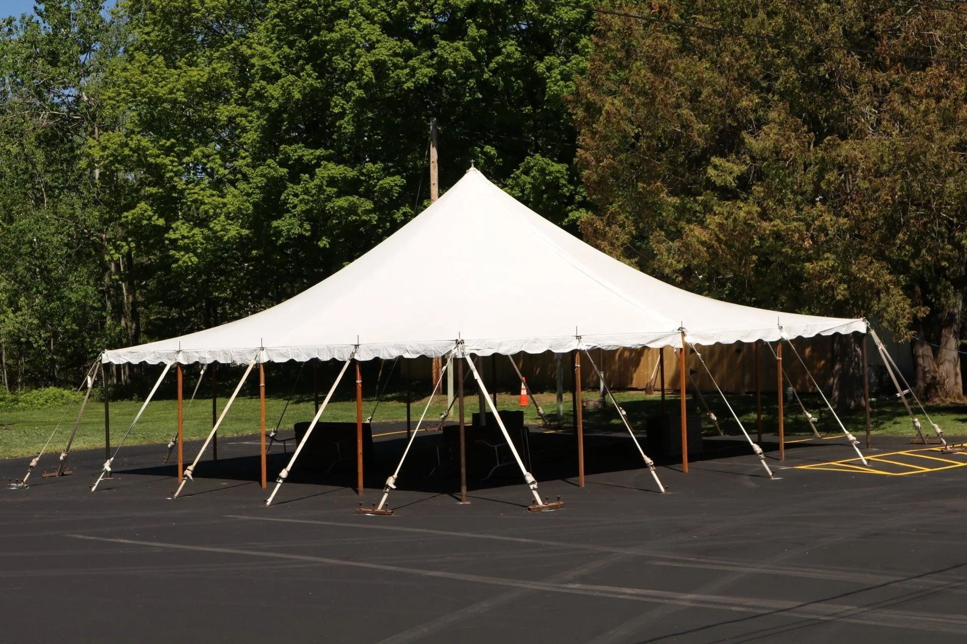 White tent with peaked roof, set up on asphalt, with trees in the background.