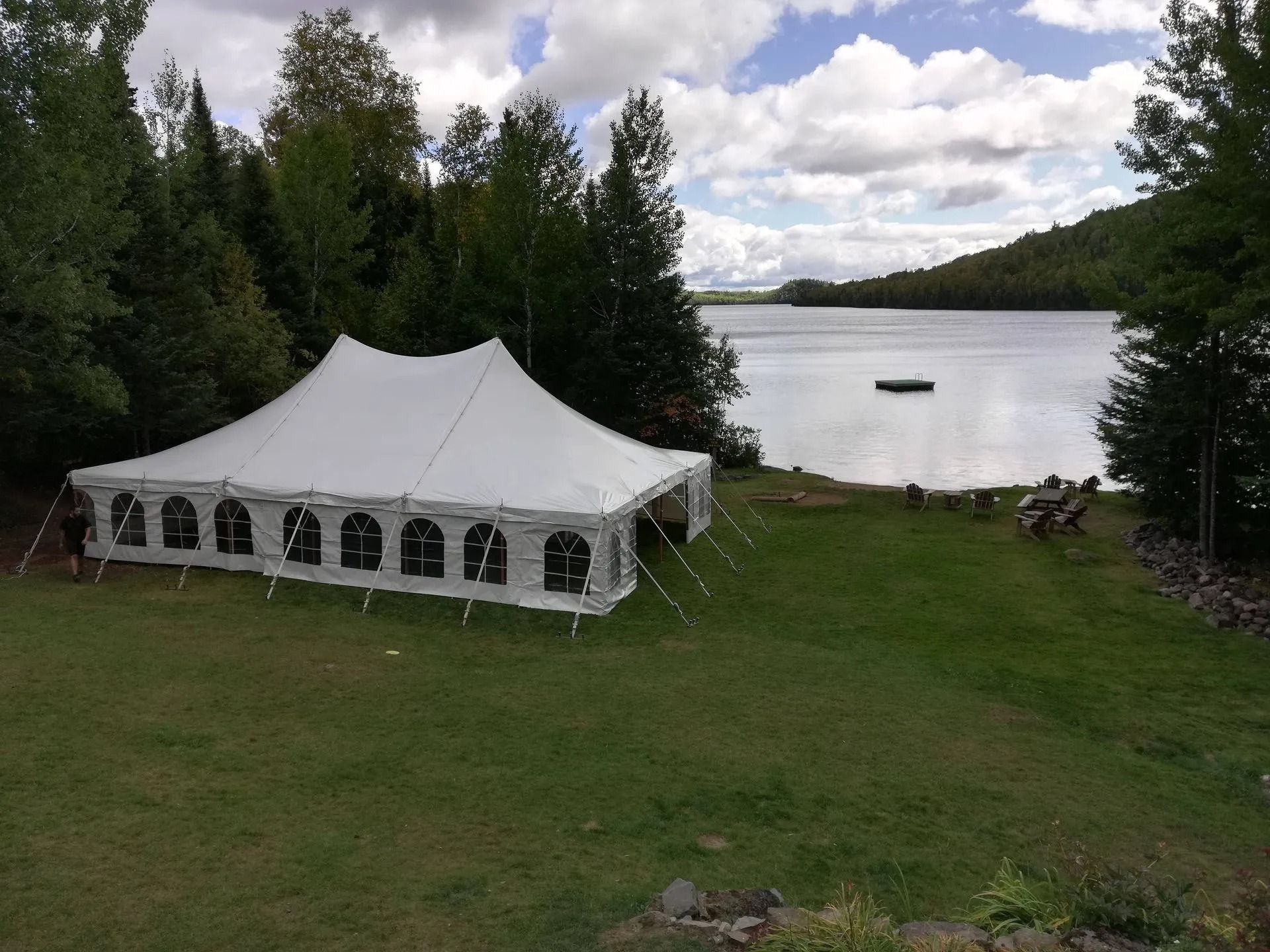 White event tent on grassy lake shore, trees in background, cloudy sky.