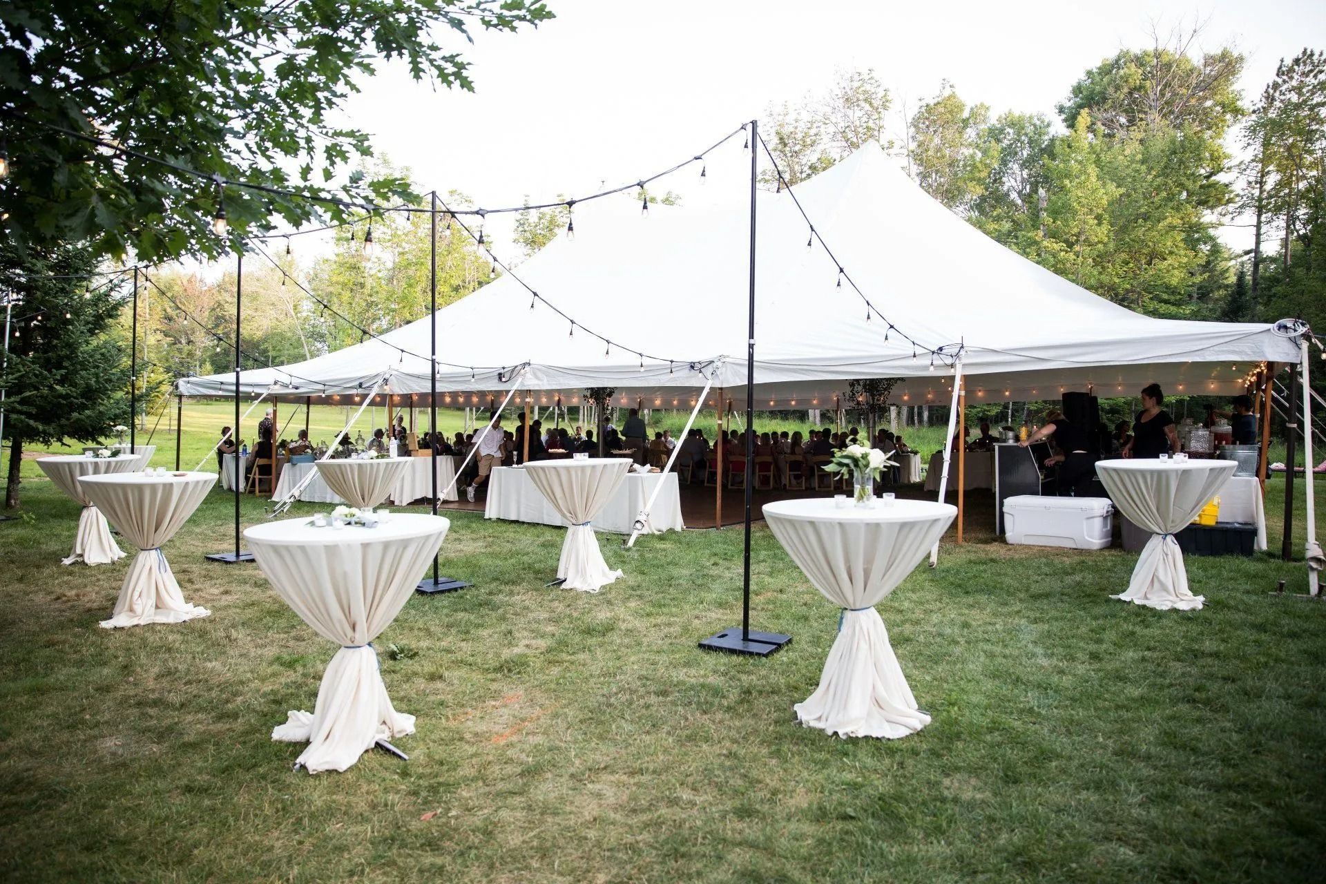 Outdoor event tent with string lights, cocktail tables, and guests in a grassy setting.