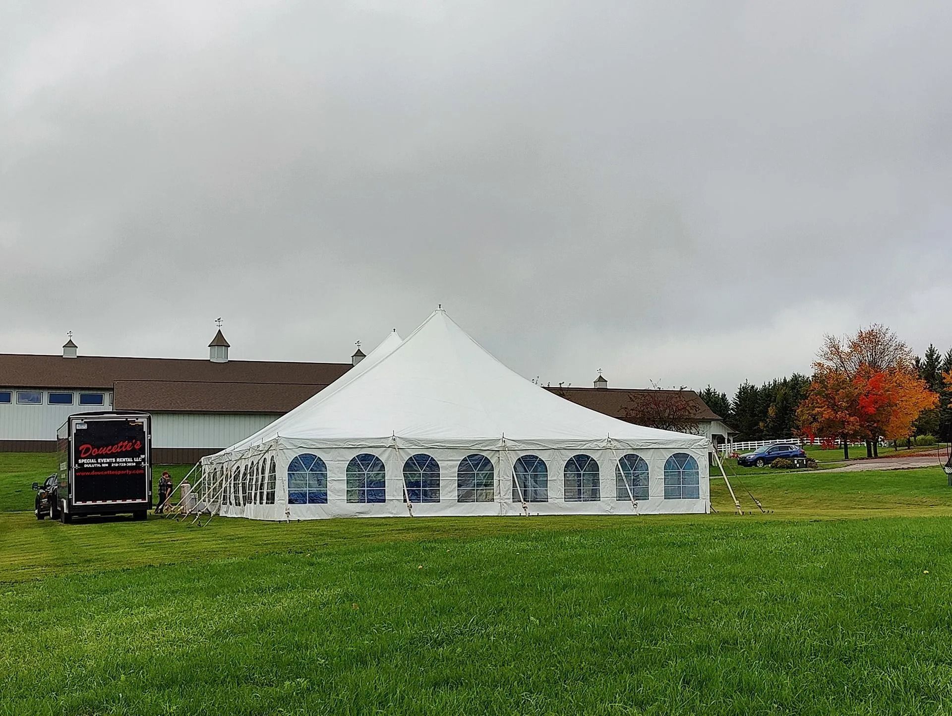 White event tent in a grassy field, with a building and colorful tree in the background. Overcast sky.