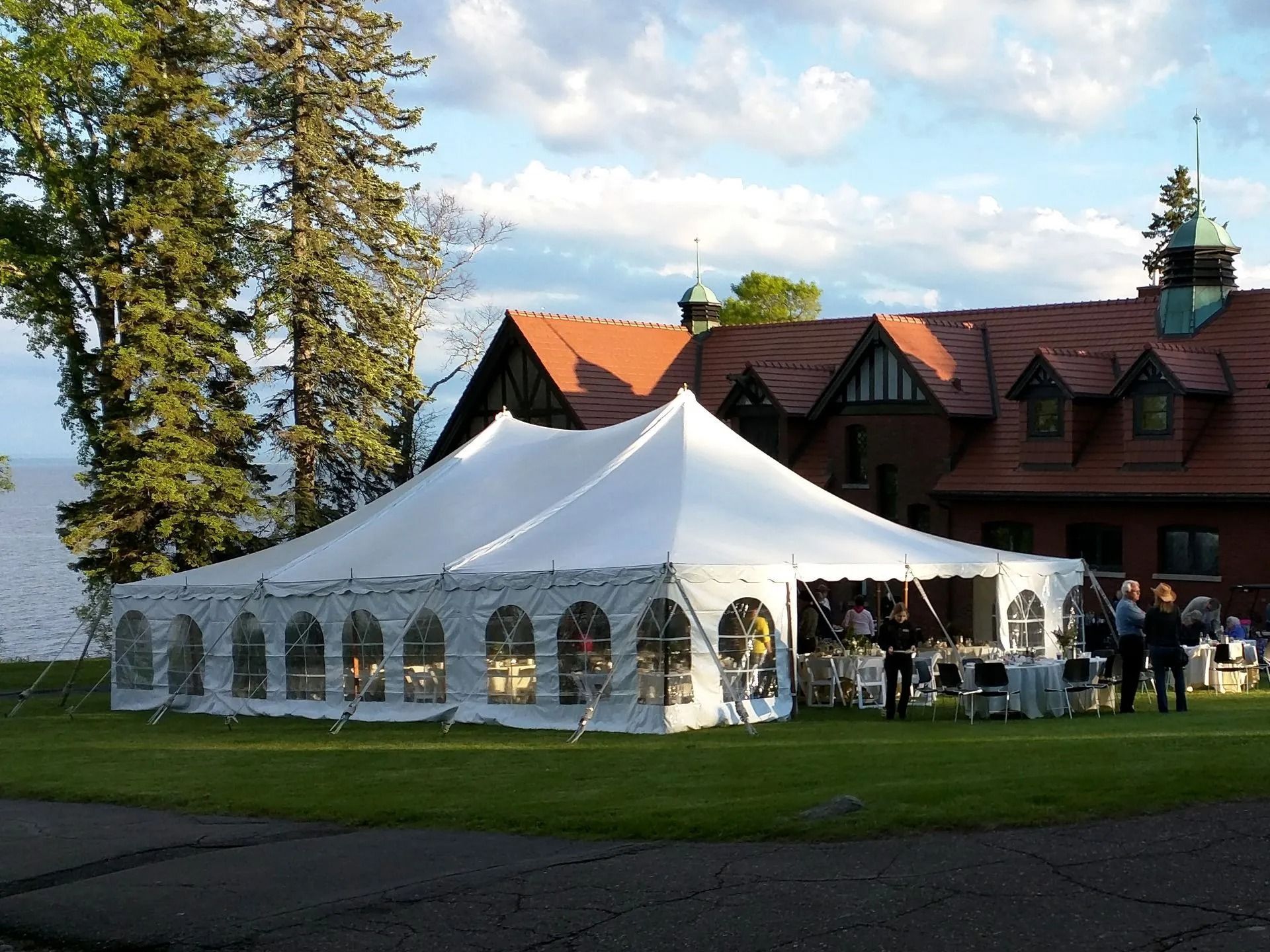 White tent set up on a lawn near a lake, next to a red-roofed building.