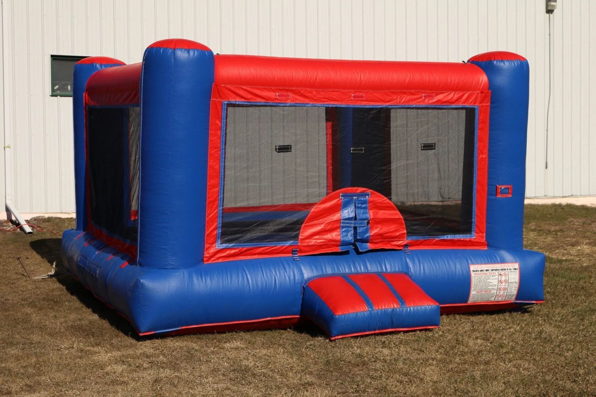 Blue and red inflatable bounce house on grass, featuring mesh windows and a step.