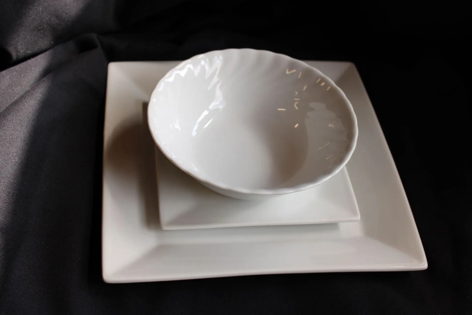 White bowl and square plates stacked on black cloth.