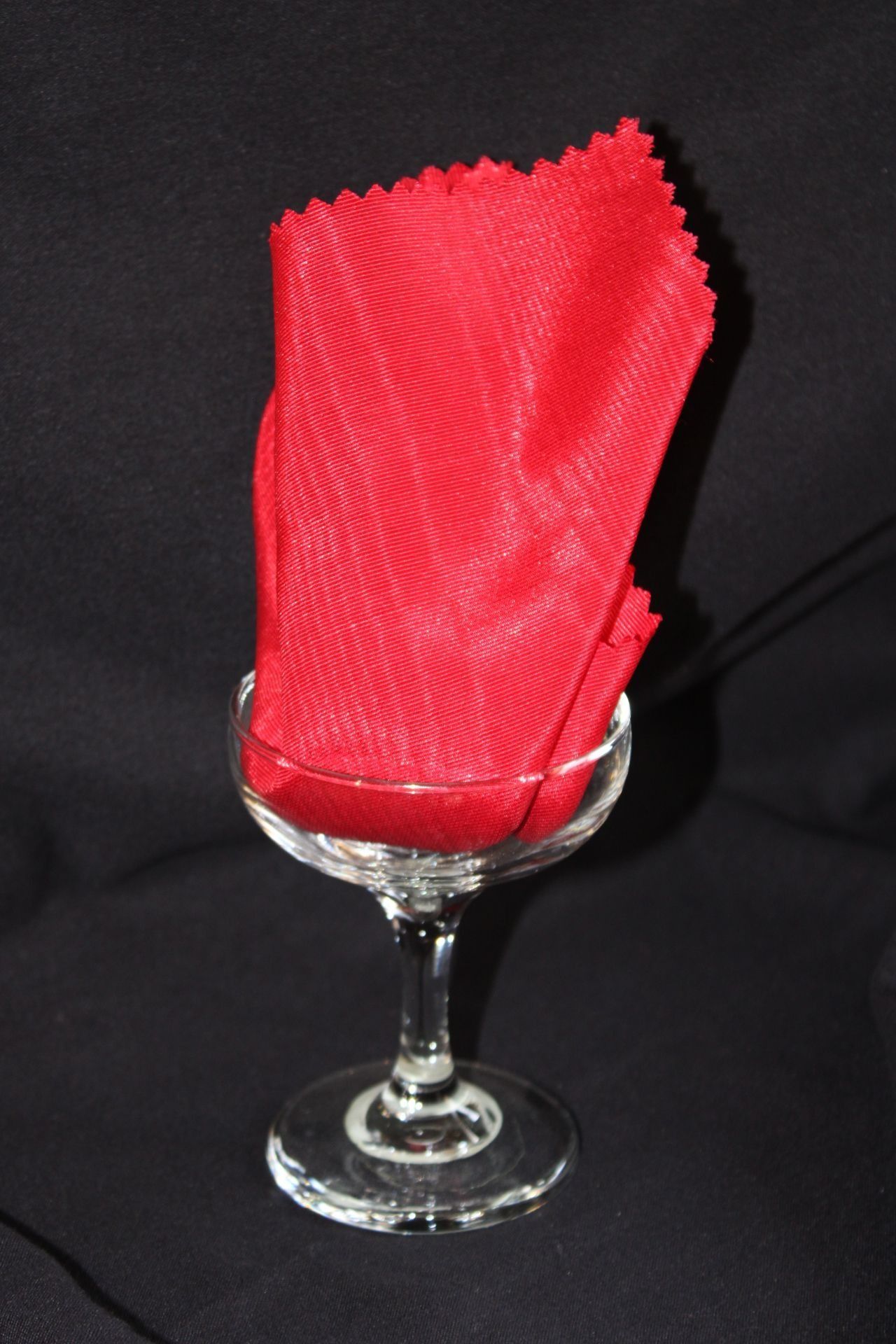 Red cloth napkin arranged in a clear glass goblet, set against a dark background.