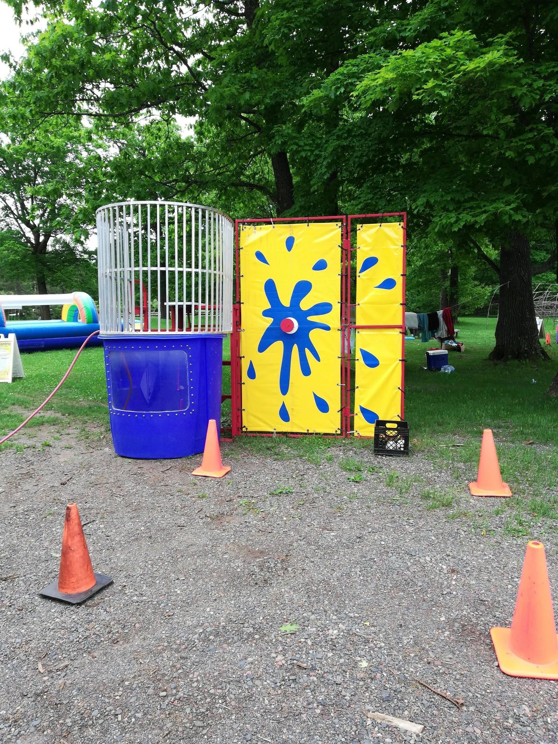 Dunk tank with a person inside, next to a yellow target on a grassy area. Cones on the ground.