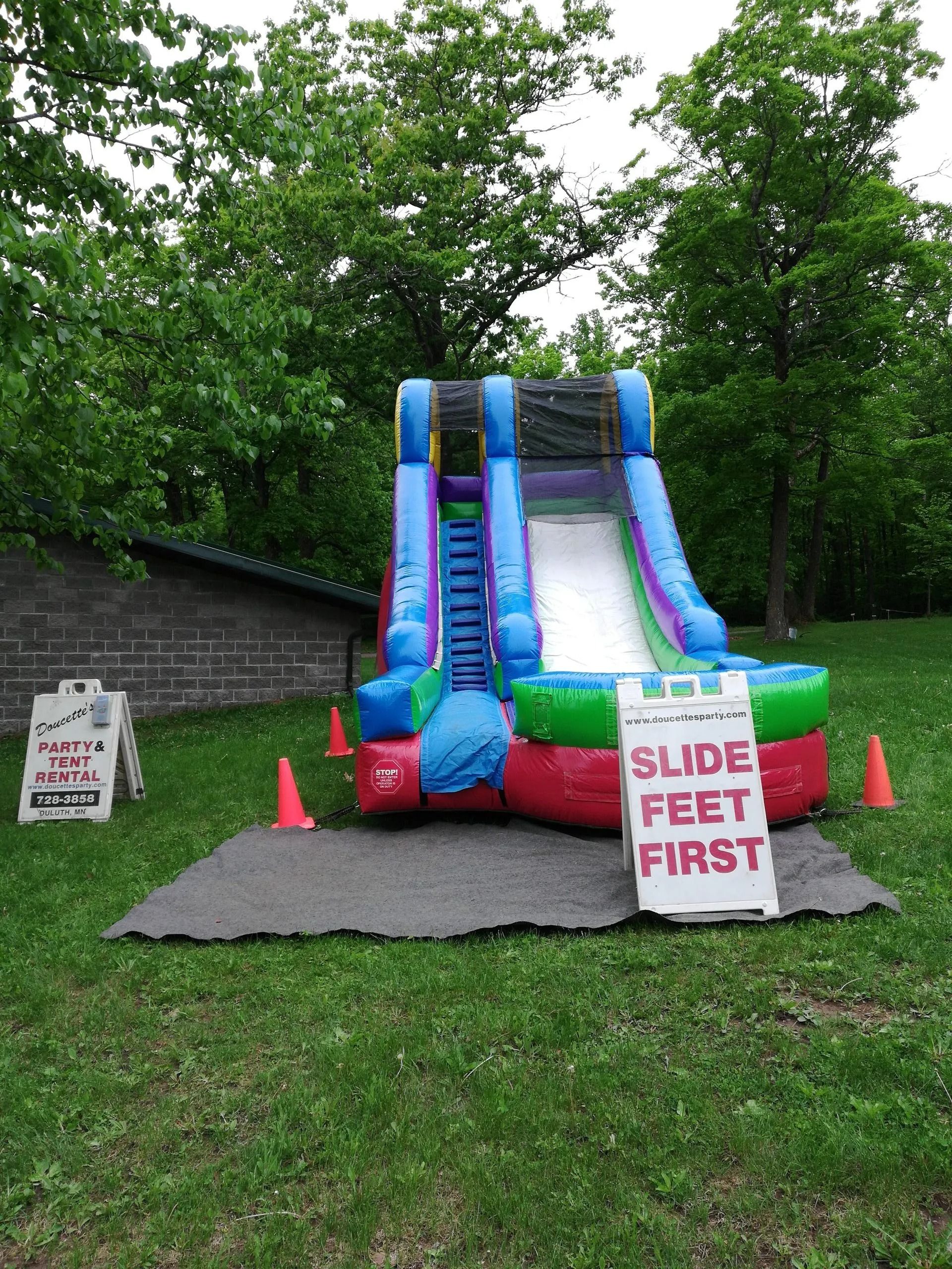 Inflatable slide on green grass with trees. Sign reads "Slide feet first."