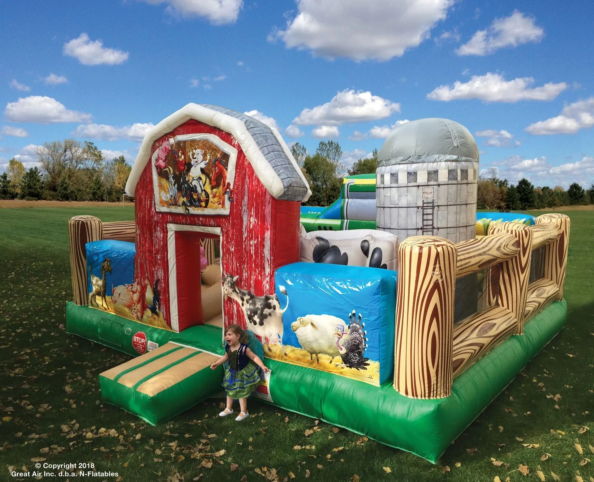 Inflatable farm-themed bounce house with red barn, silo, and child entering. Green field, blue sky.