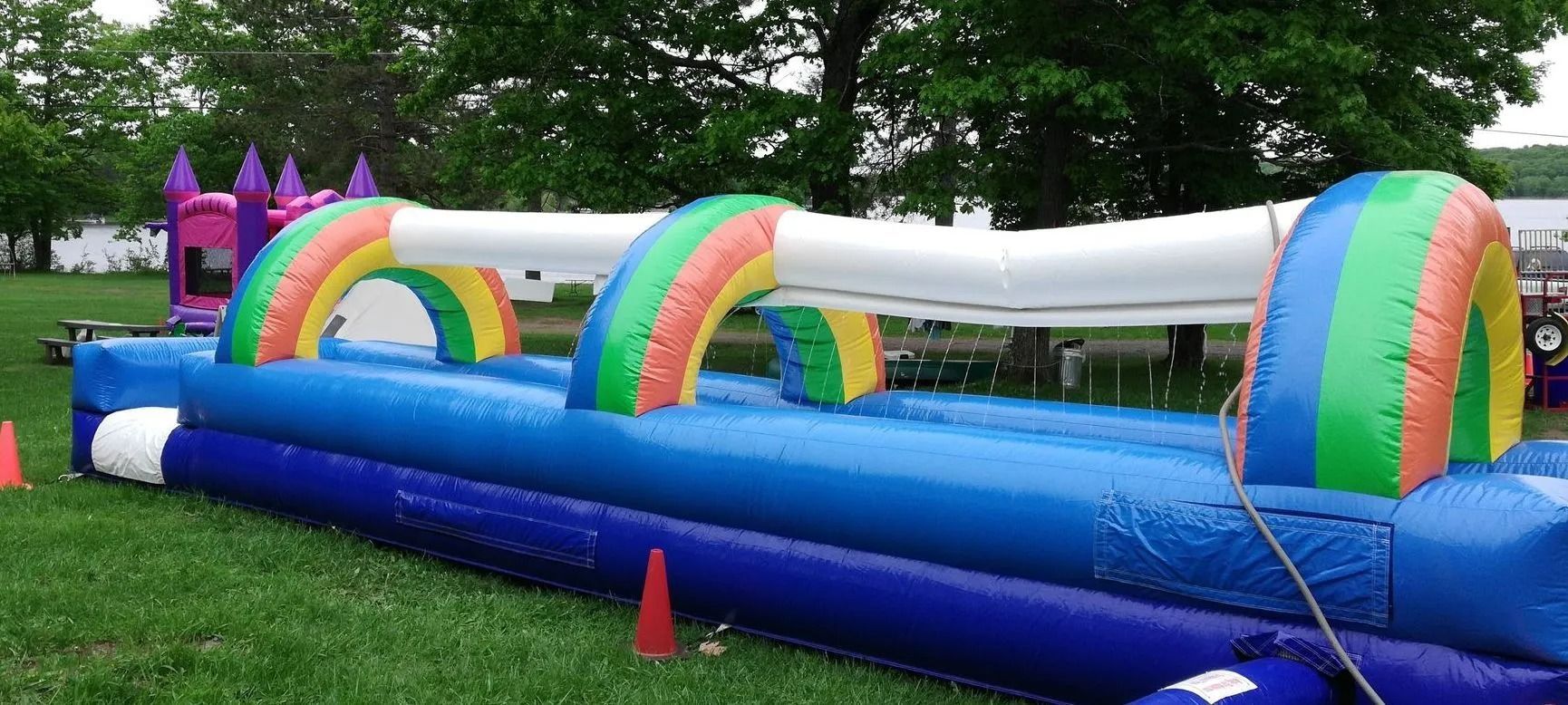 A large, inflatable water slide with rainbow arches on a green lawn. There is a bounce house visible in the background.
