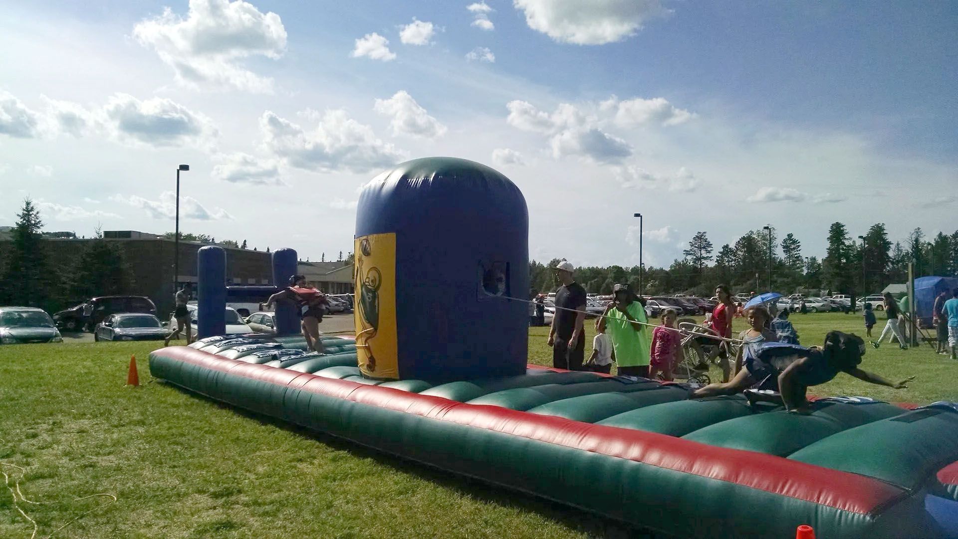 People playing on an inflatable obstacle course outdoors. Green grass, blue sky, and cars.