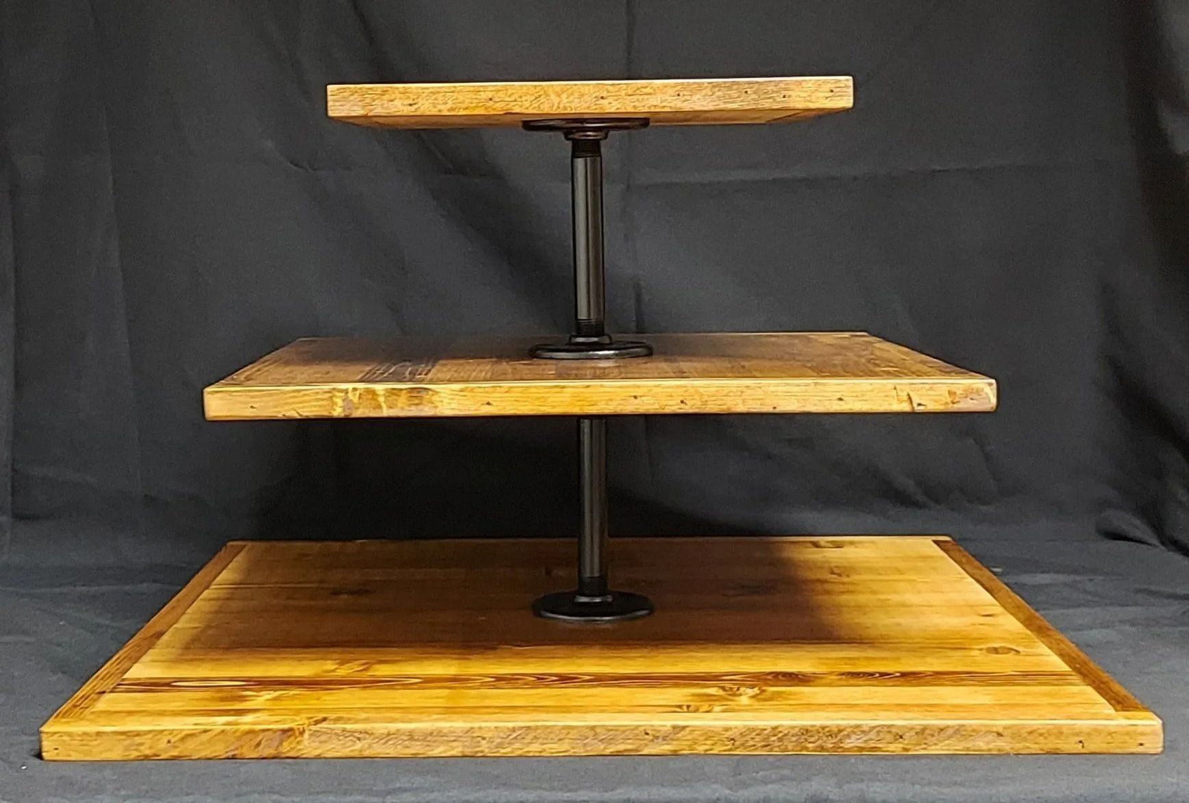 Three-tiered wooden stand with a rustic finish, supported by black metal pipes, against a black backdrop.