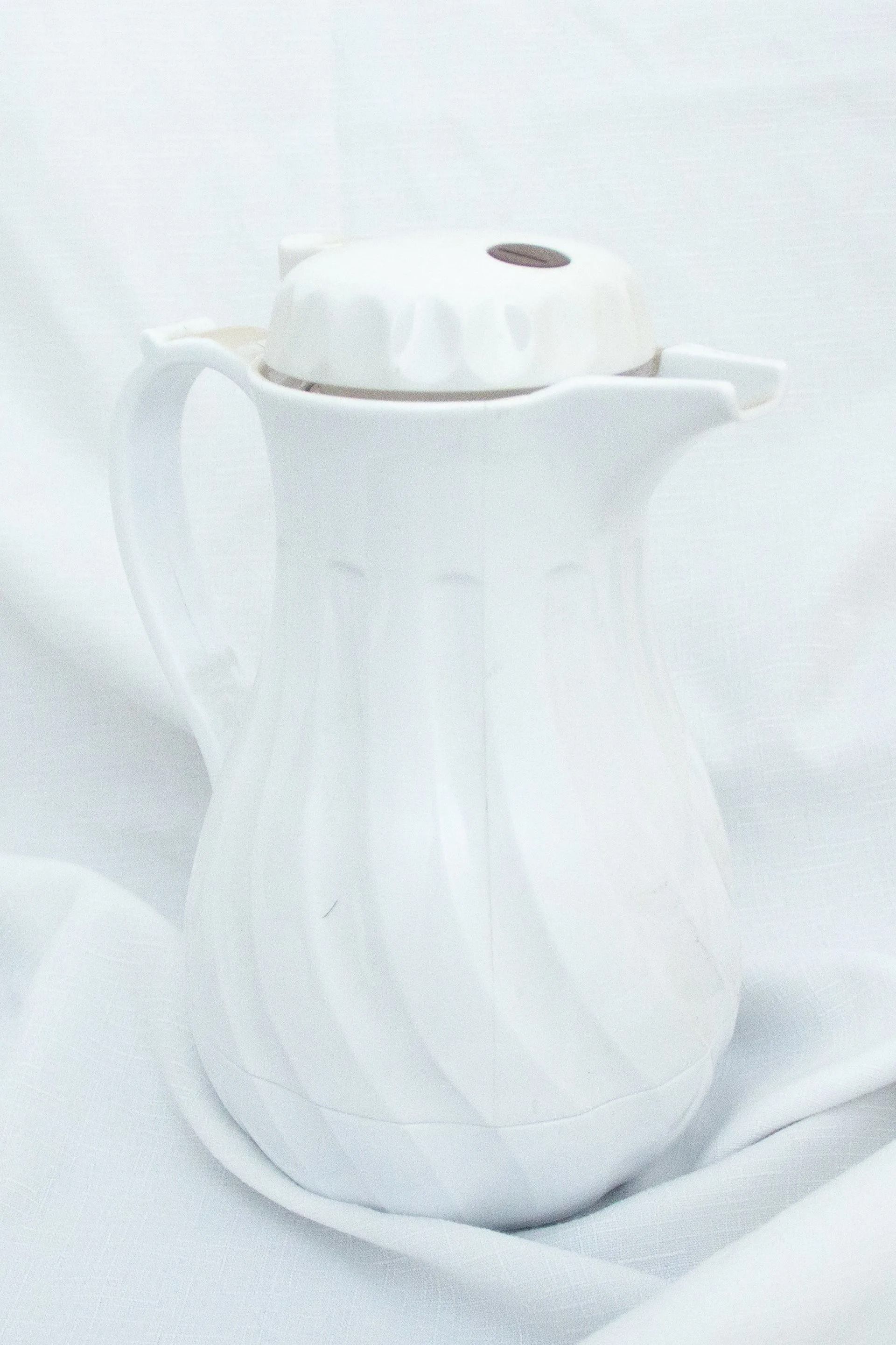 White ceramic pitcher with a fluted design and matching lid, resting on white fabric.