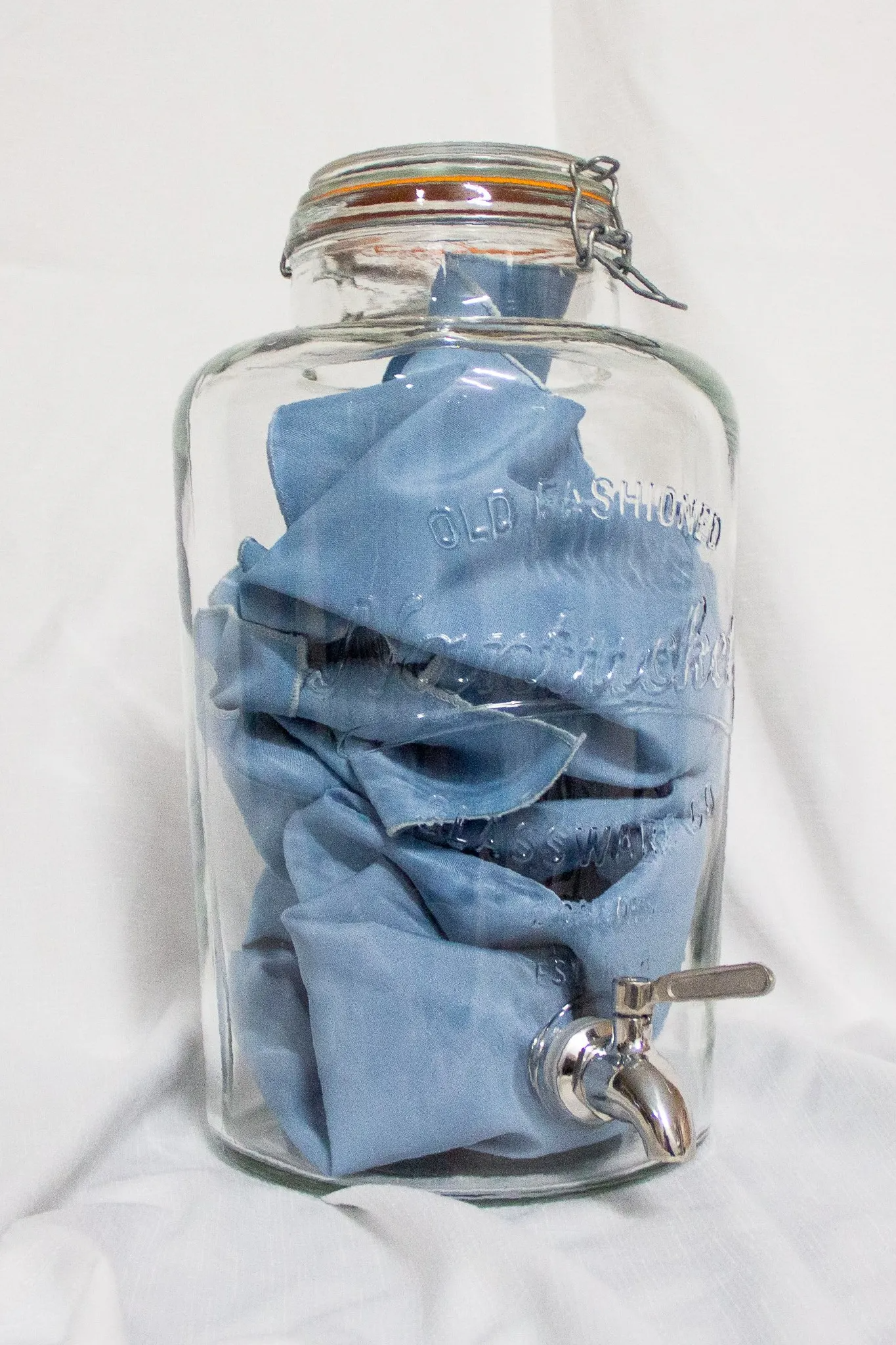 Clear glass beverage dispenser with blue cloth inside; silver spigot and latch.
