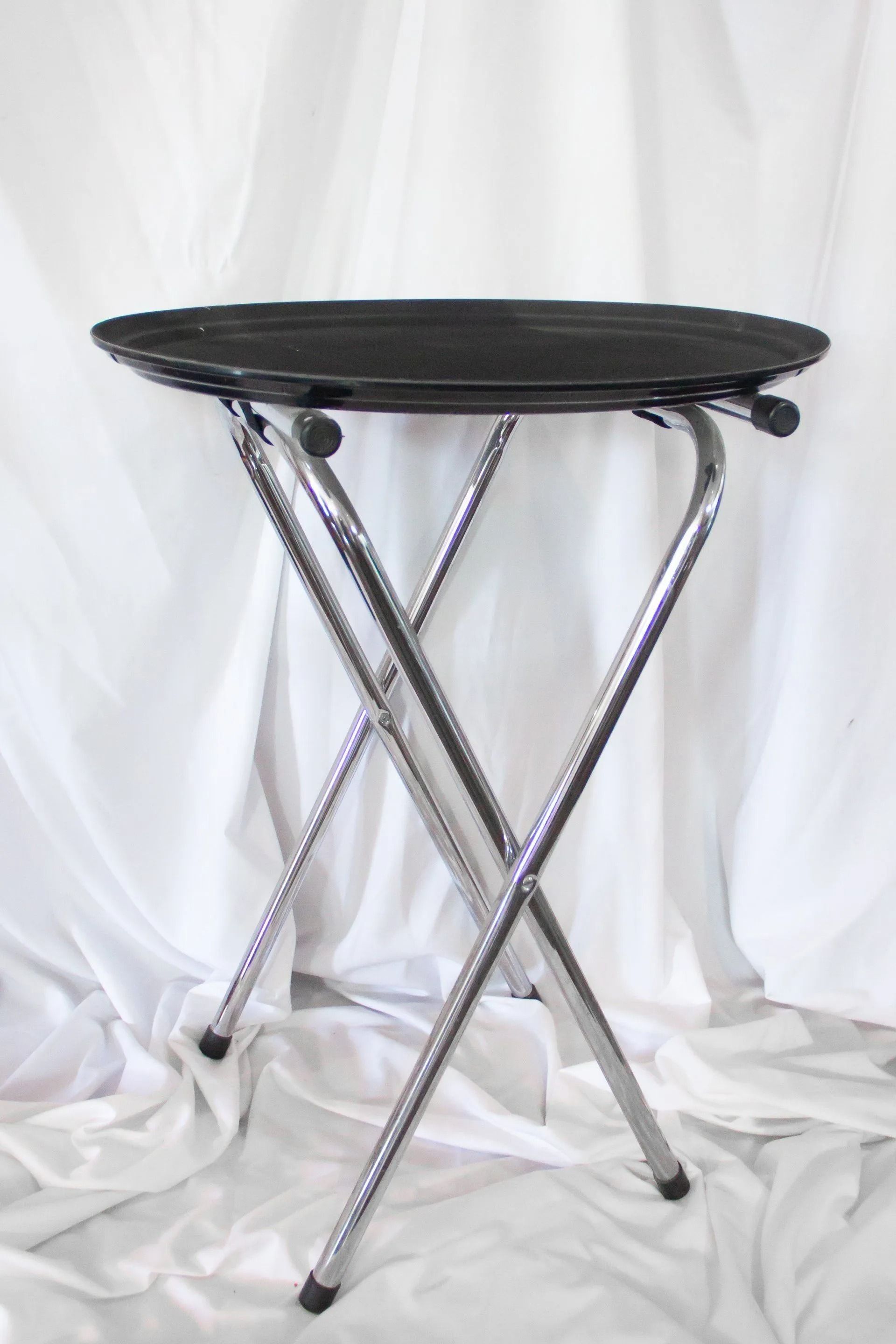 Round black tray table, chrome folding base. White backdrop.