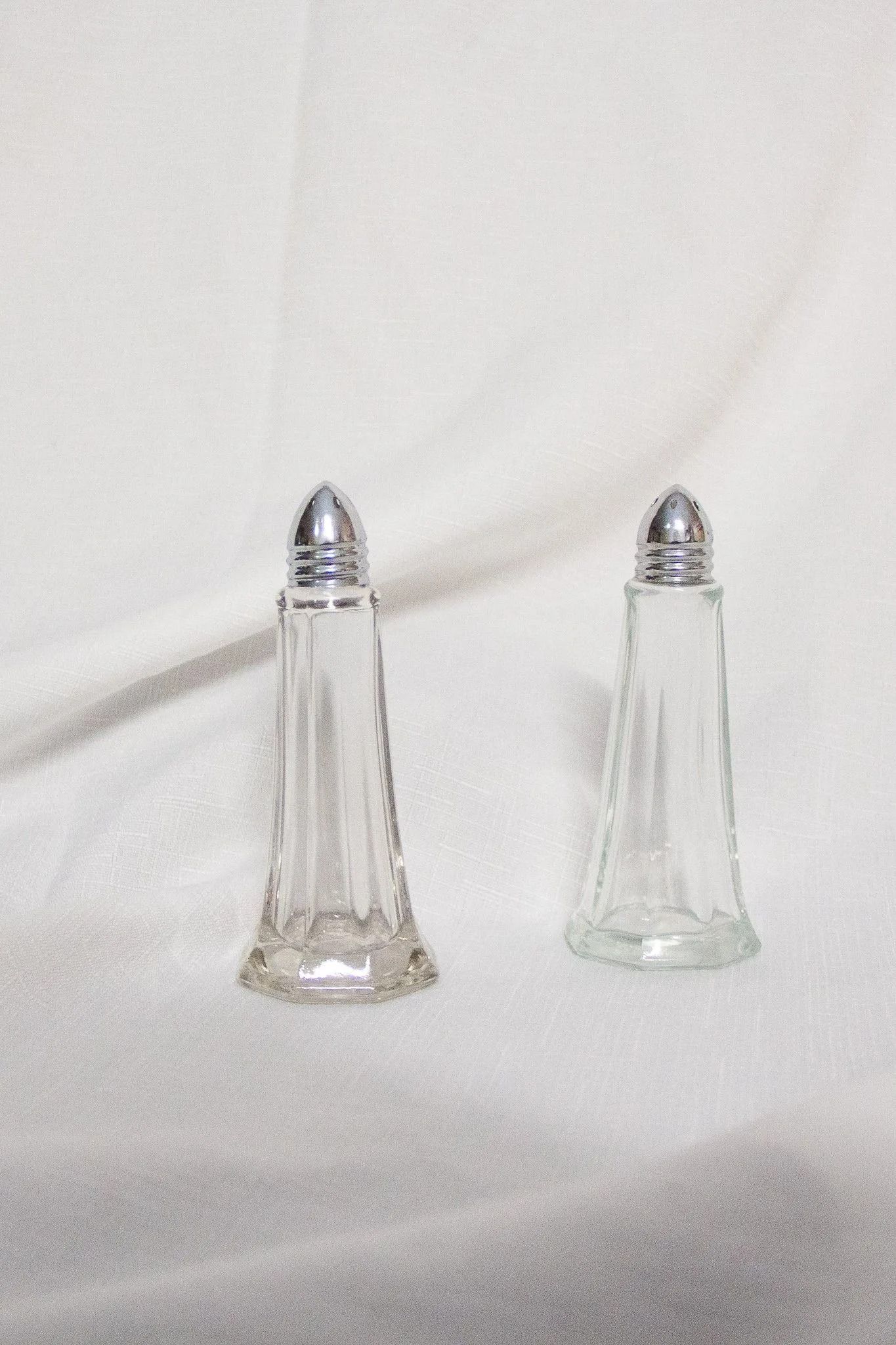 Two clear glass salt shakers with silver tops, on a white textured surface.