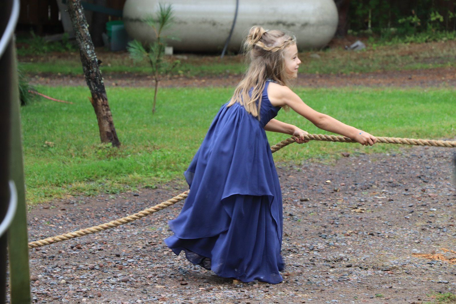 Girl in blue dress pulls on a rope outdoors; gravel path, green grass, and white tank in background.