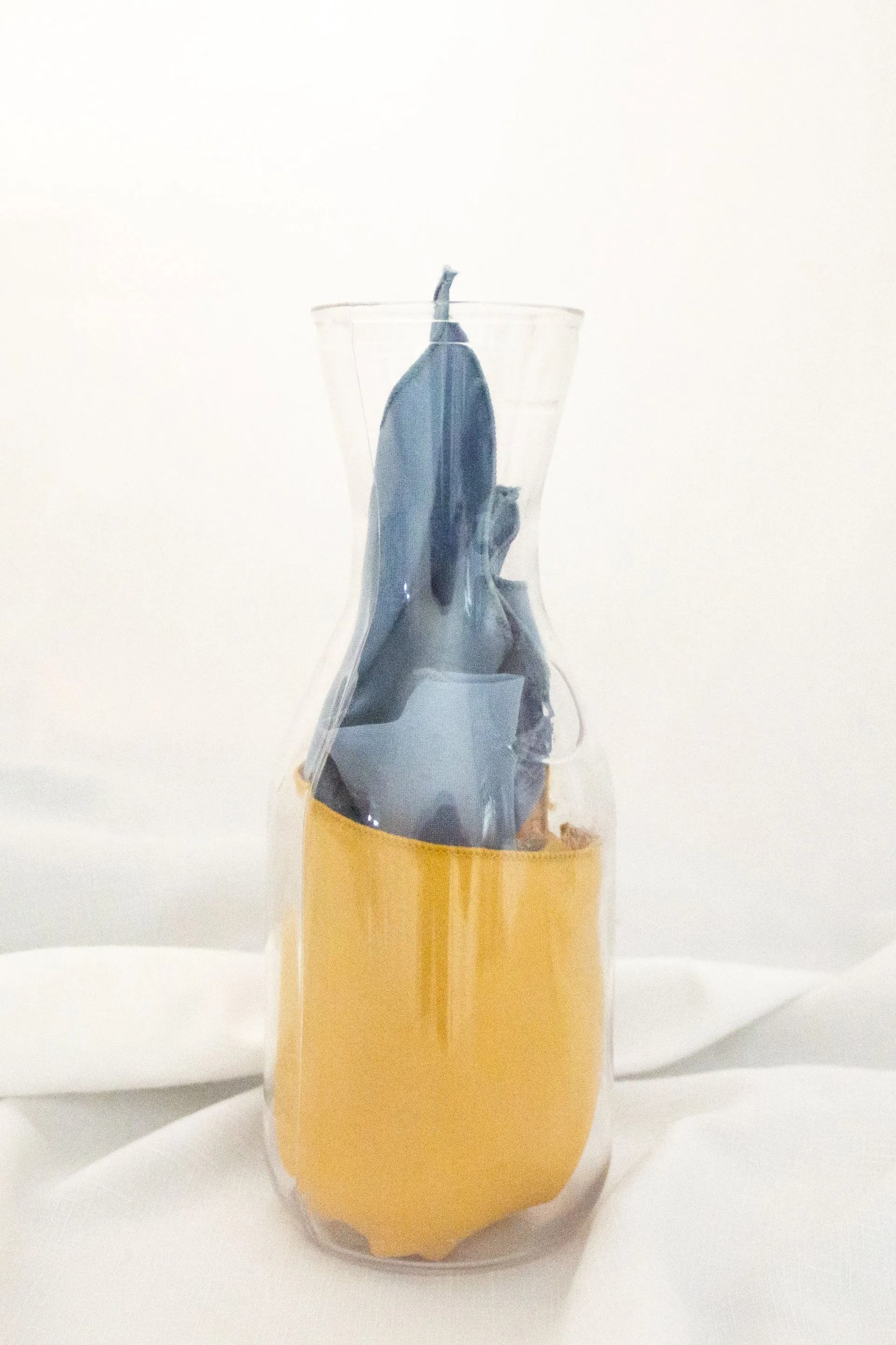 Blue candle in clear hourglass-shaped vase, yellow sand base, white cloth background.