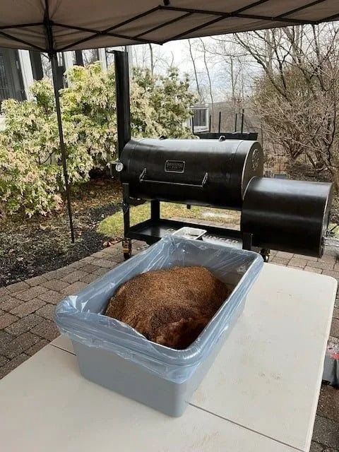Smoked meat in a bin on a table, with a smoker grill in the background, outdoors.