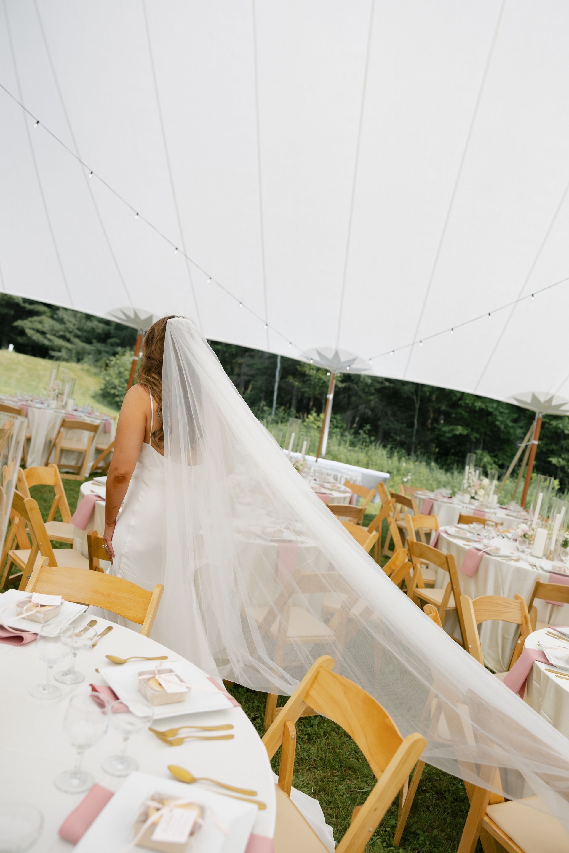 Bride in a wedding dress, walking near decorated tables with a long veil trailing.