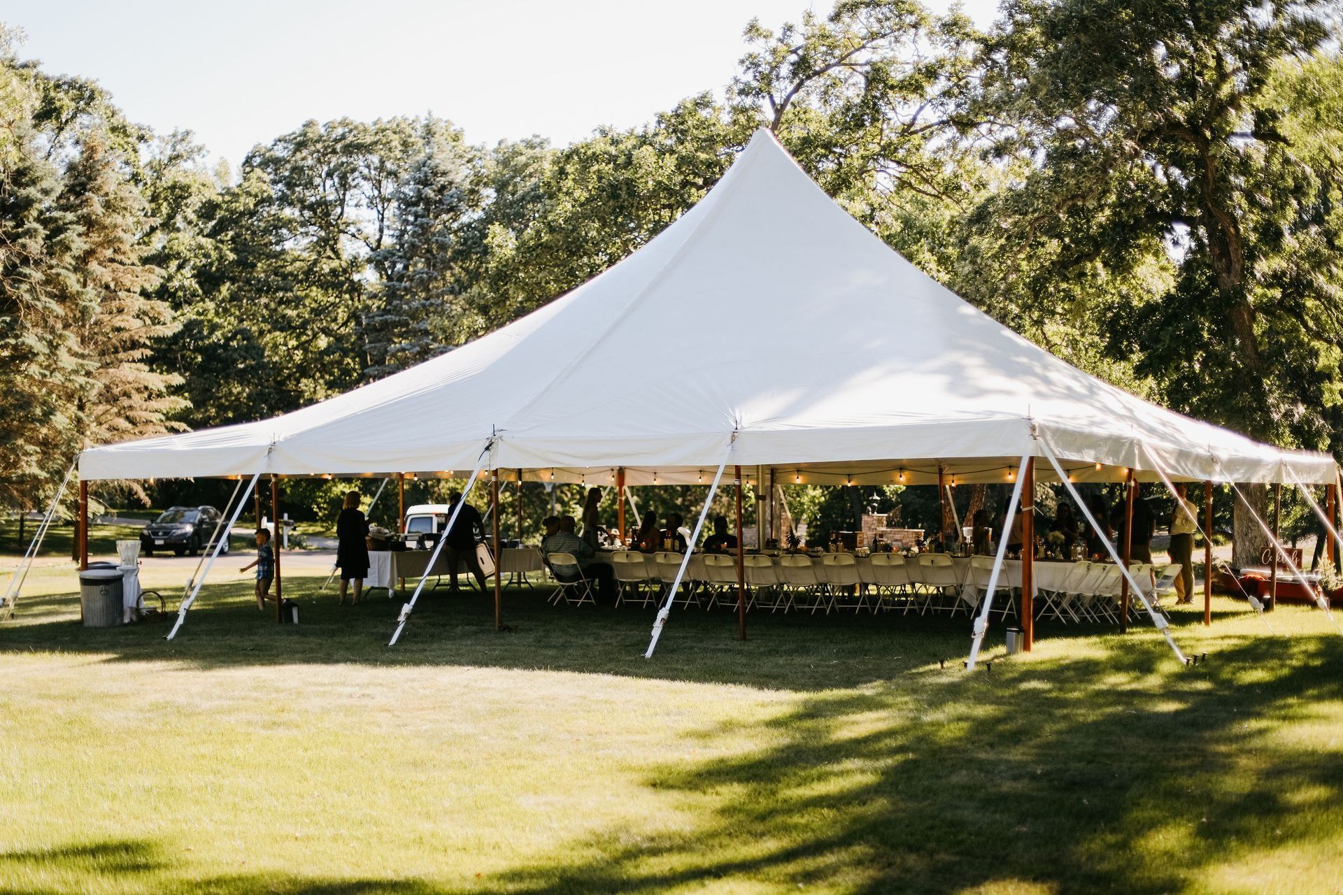 White tent set up on grassy lawn; people and tables inside, trees in background.