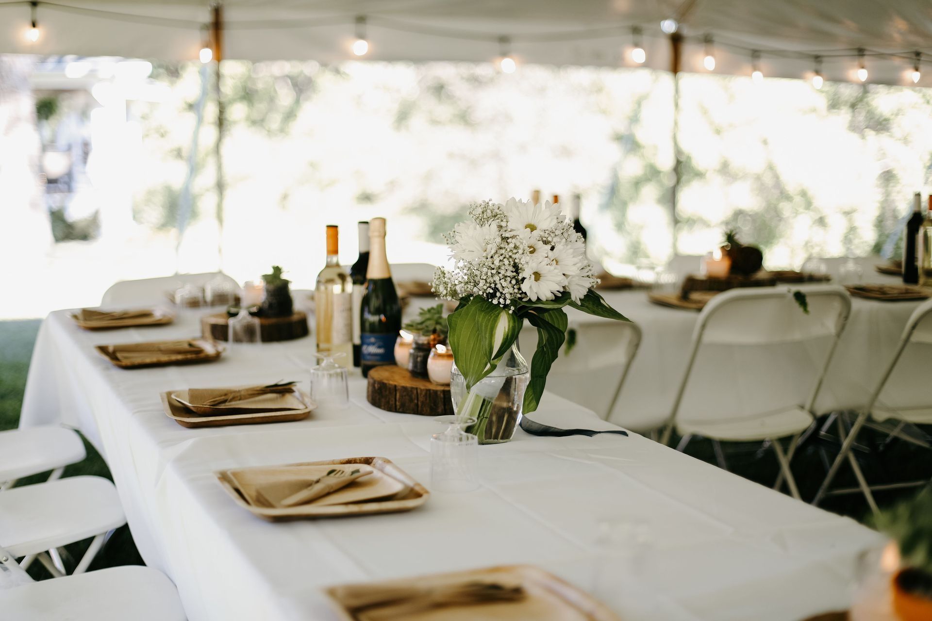 Table set for an outdoor event, white tablecloth, floral centerpiece, plates, utensils, and bottles.