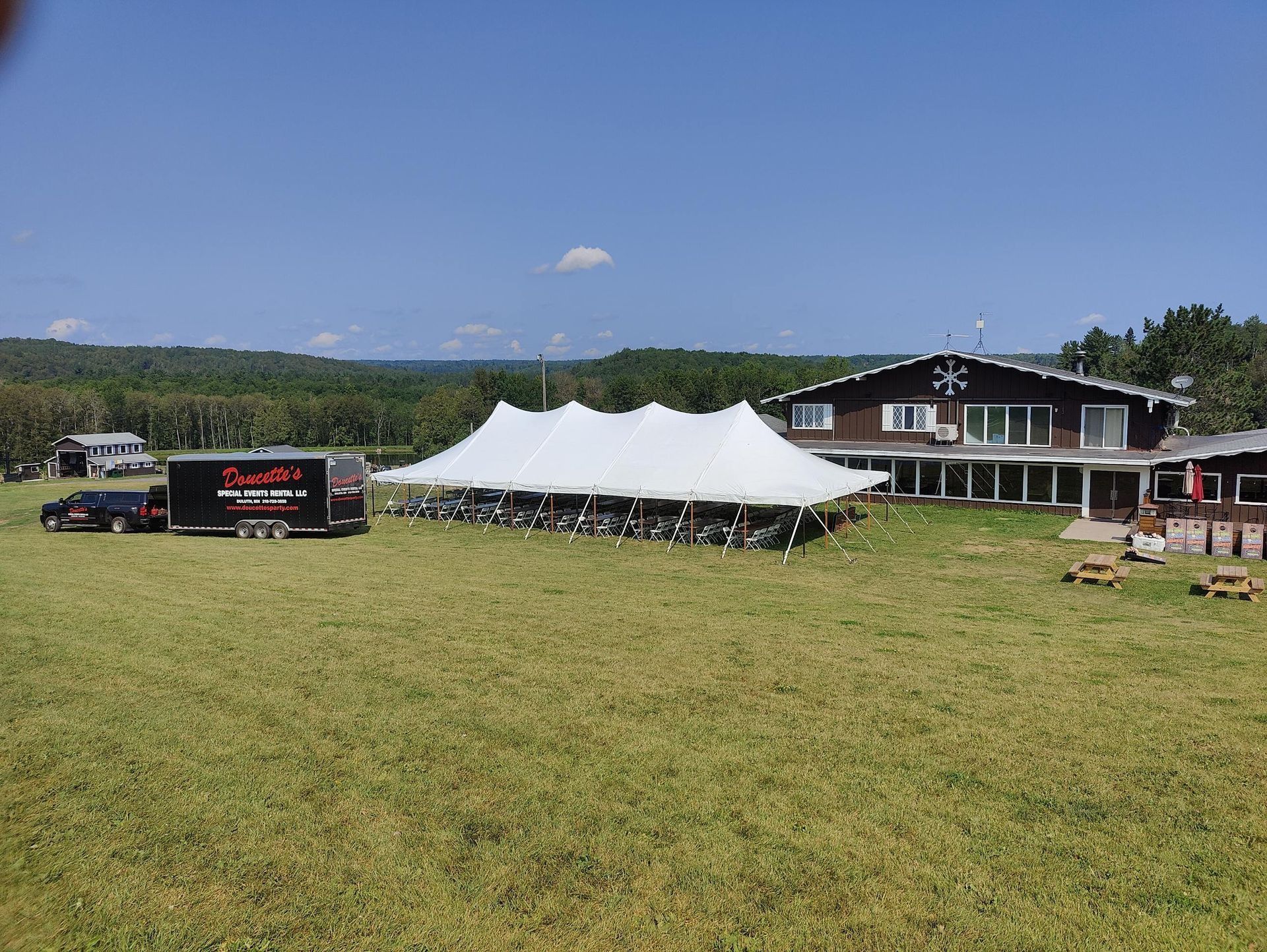 White event tent on a grassy field, near a lodge-style building. Black trailer parked nearby. Sunny day.