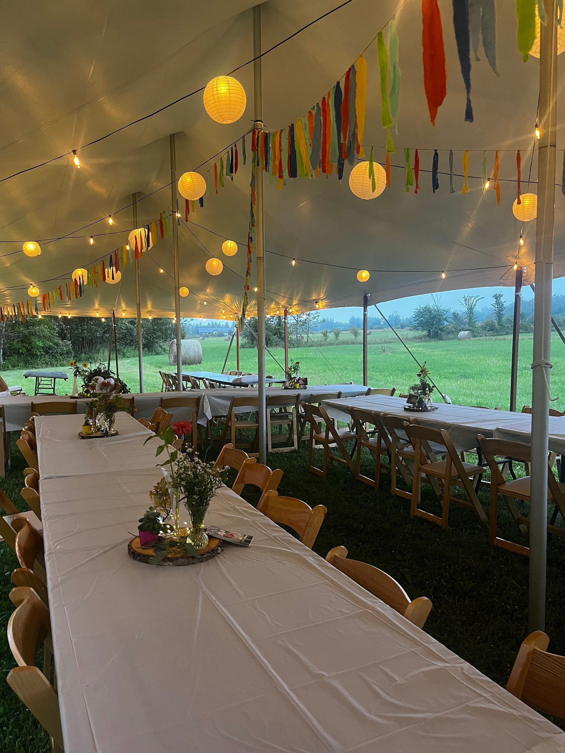 Long tables set for a gathering under a tent with string lights and colorful decorations, in a grassy field.