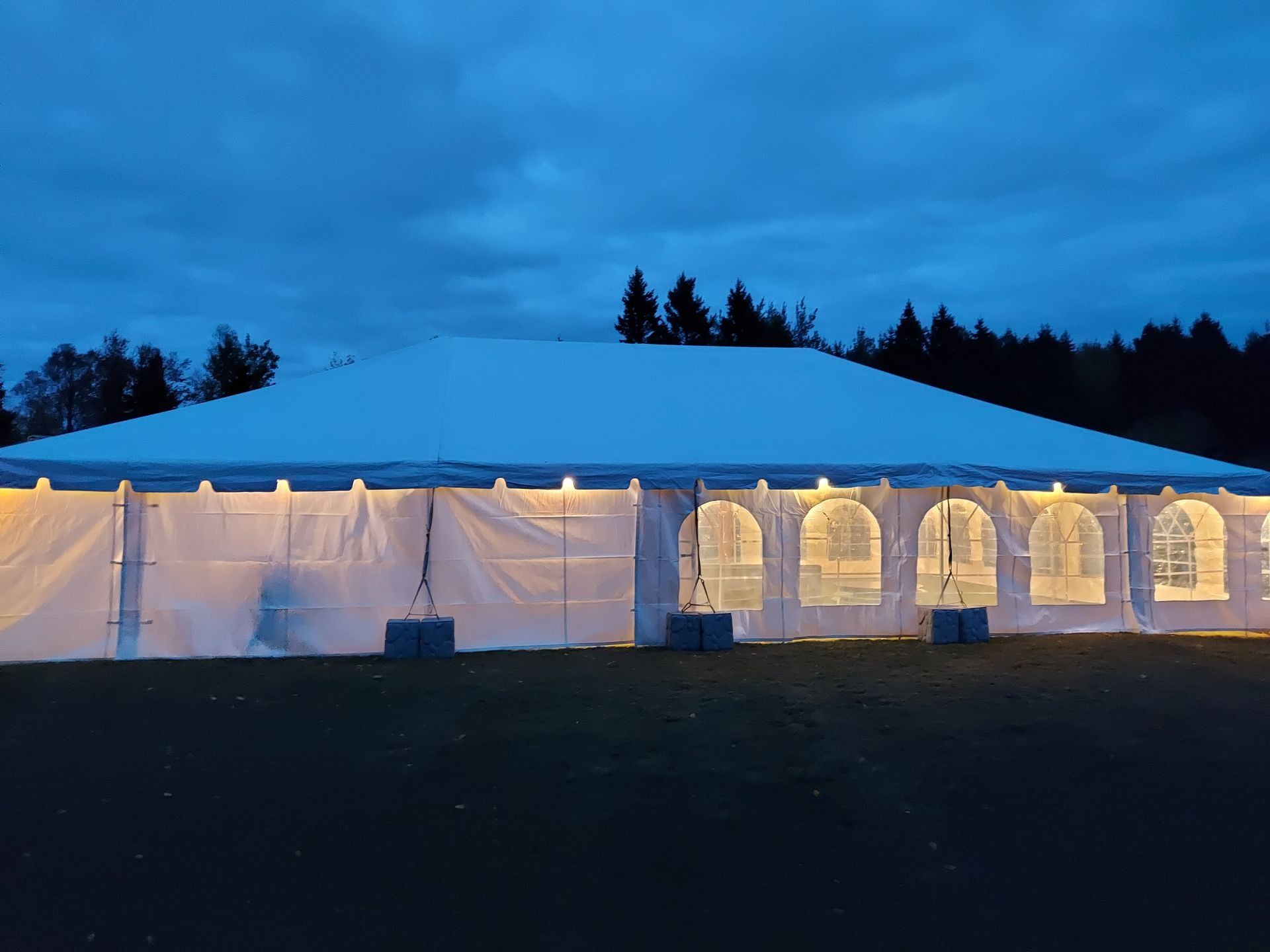 White tent with lighted windows against a dusk sky and trees.