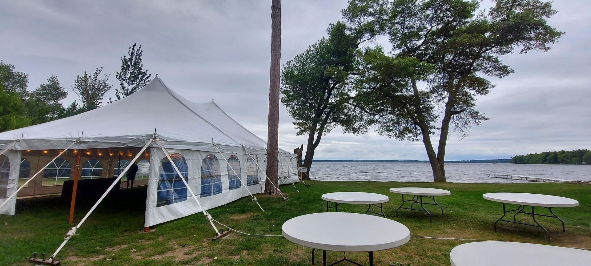A white tent set up on a grassy area next to a lake with round white tables.