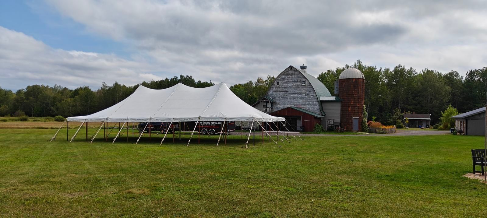 White tent set up on a grassy field in front of a barn and silo under a cloudy sky.