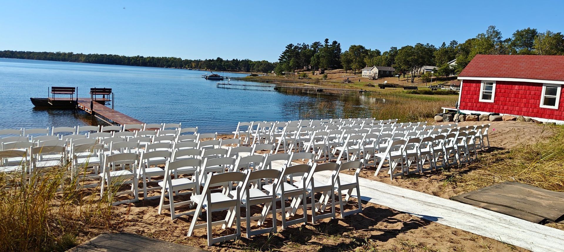 Beach wedding venue with rows of white chairs facing a lake and a red building.