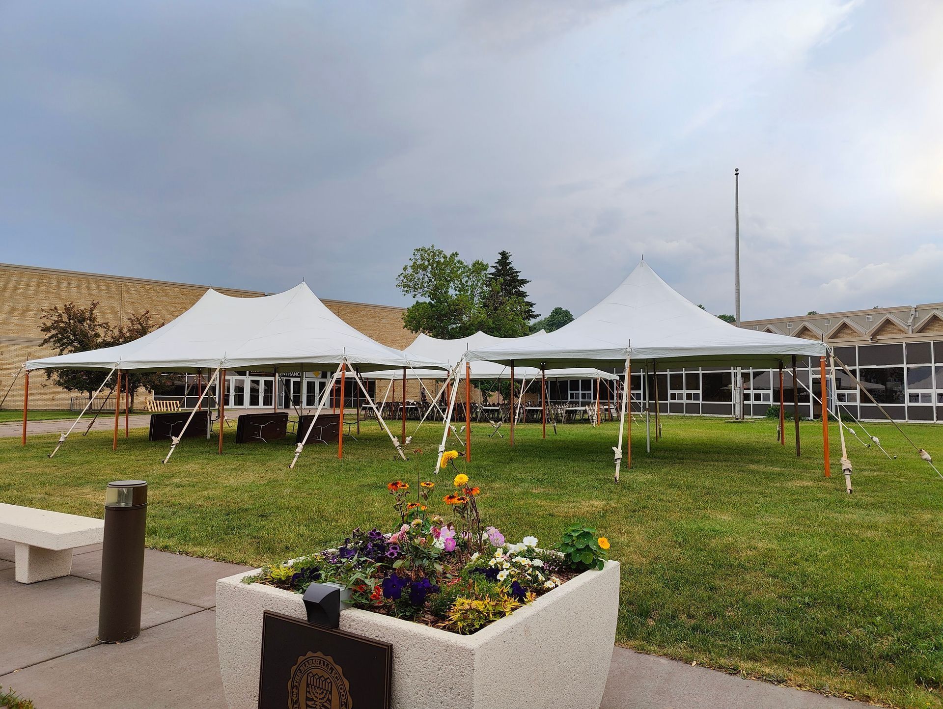 White tents set up on a green lawn with flowers in front of a building. Cloudy sky overhead.
