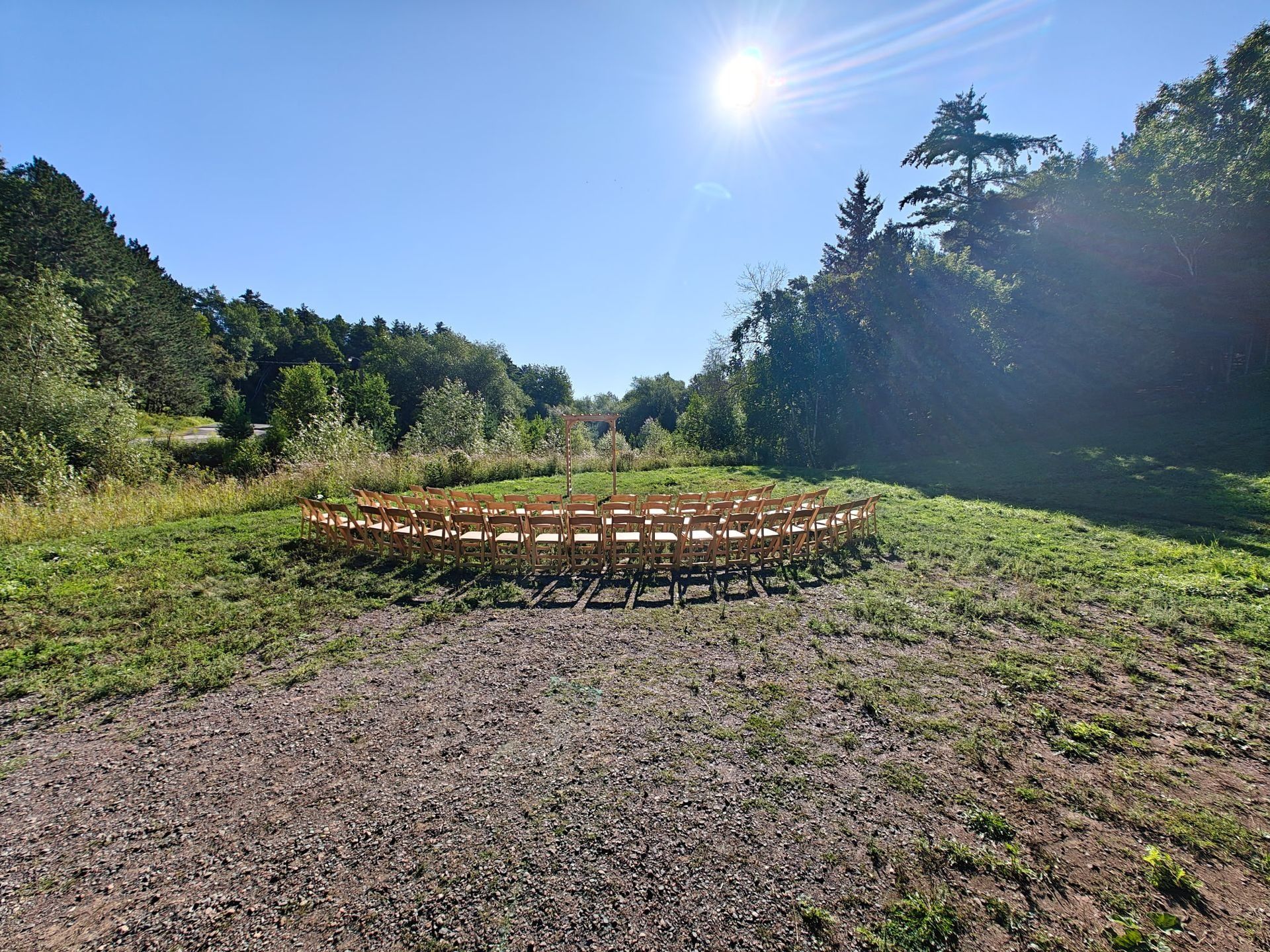 A semi-circle of wooden chairs sits on grass in a clearing, under a bright sun. Trees border the site.