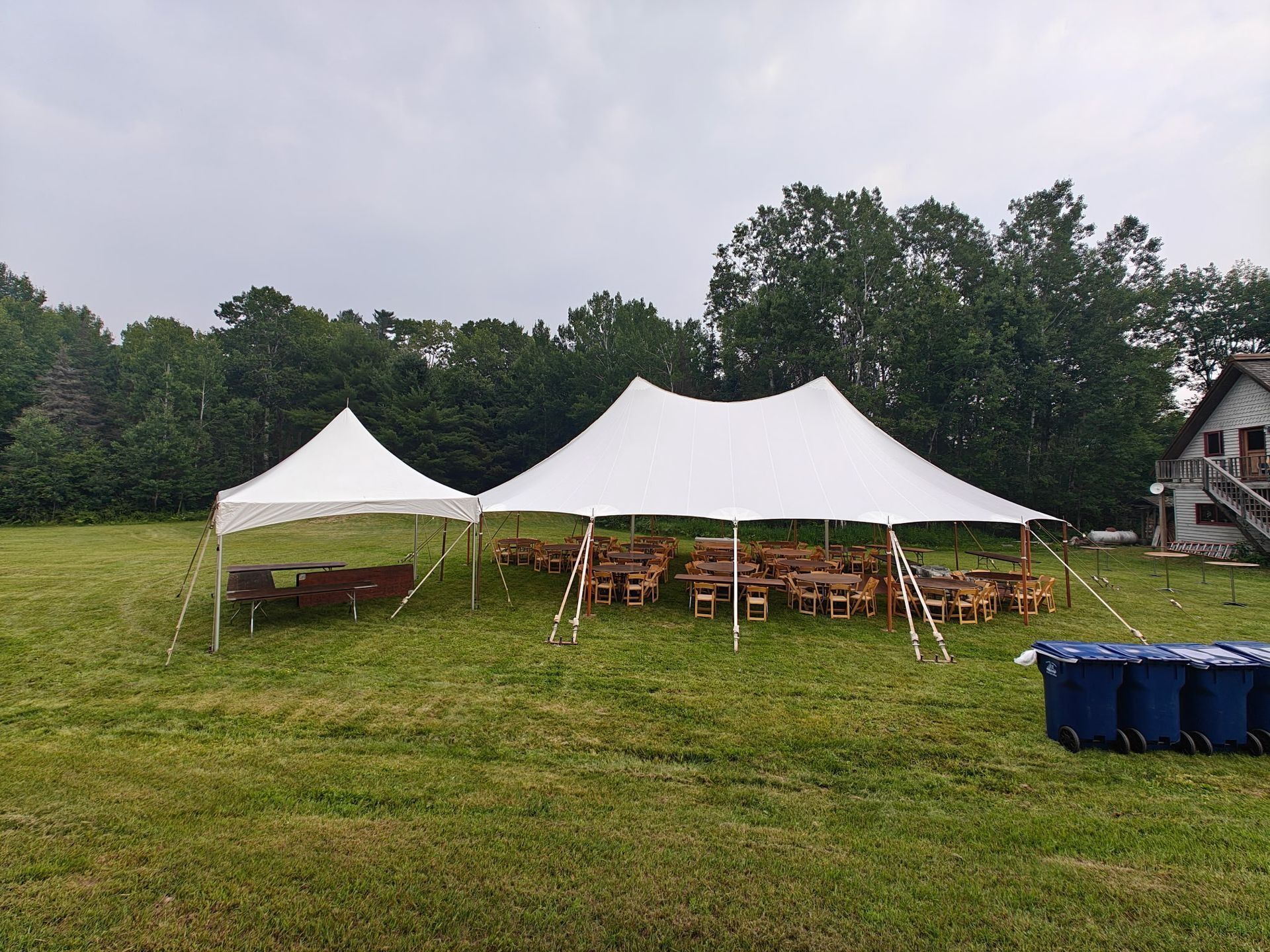 White tents set up in a grassy field with tables and chairs, trees in the background, blue bins to the right.