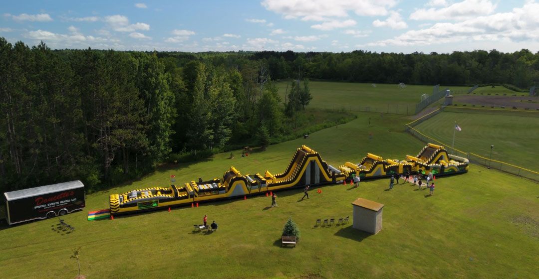 An inflatable obstacle course on green grass with people, near a forest and small structures.