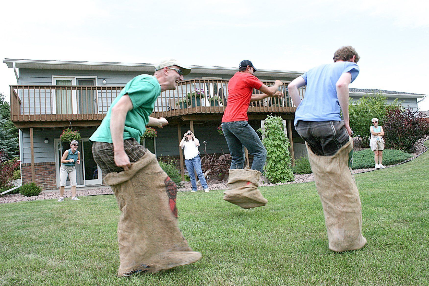 Three people in burlap sacks racing on a grassy lawn in front of a house.