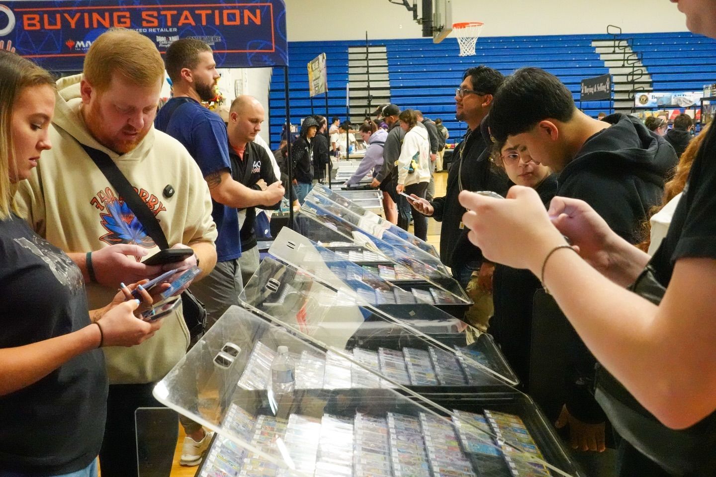 People browsing collectible cards at a buying station in a gymnasium, with rows of display cases.